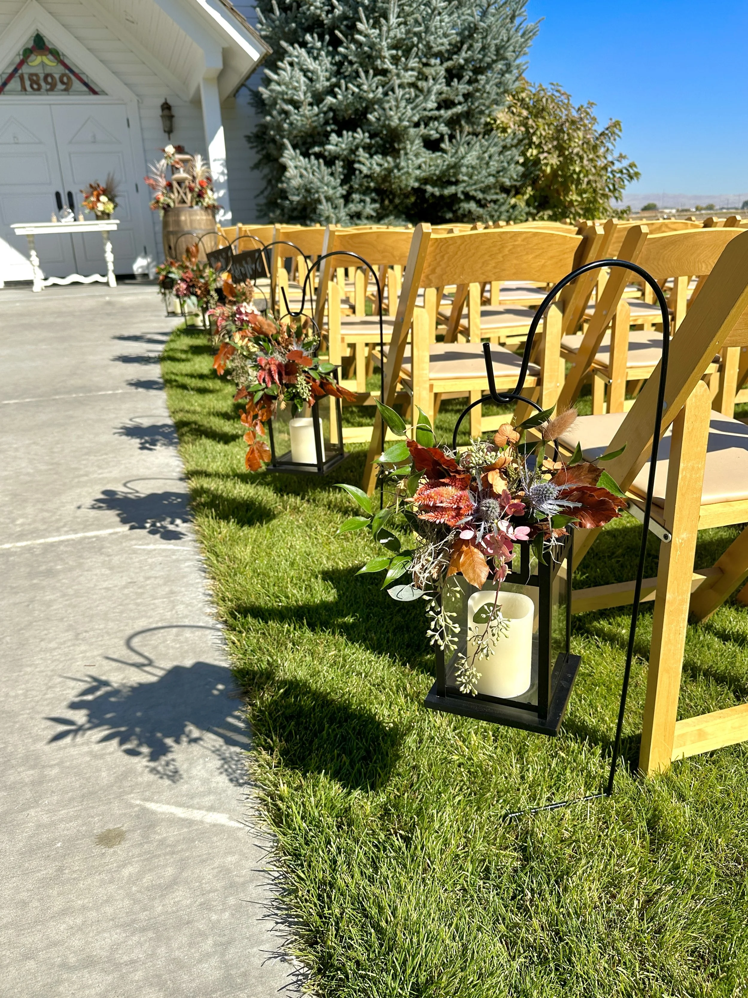 Outdoor wedding seating area decorated with autumn-themed floral arrangements in lanterns along a grassy walkway. Wedding flowers by Boise florist Edwards Flowershop.