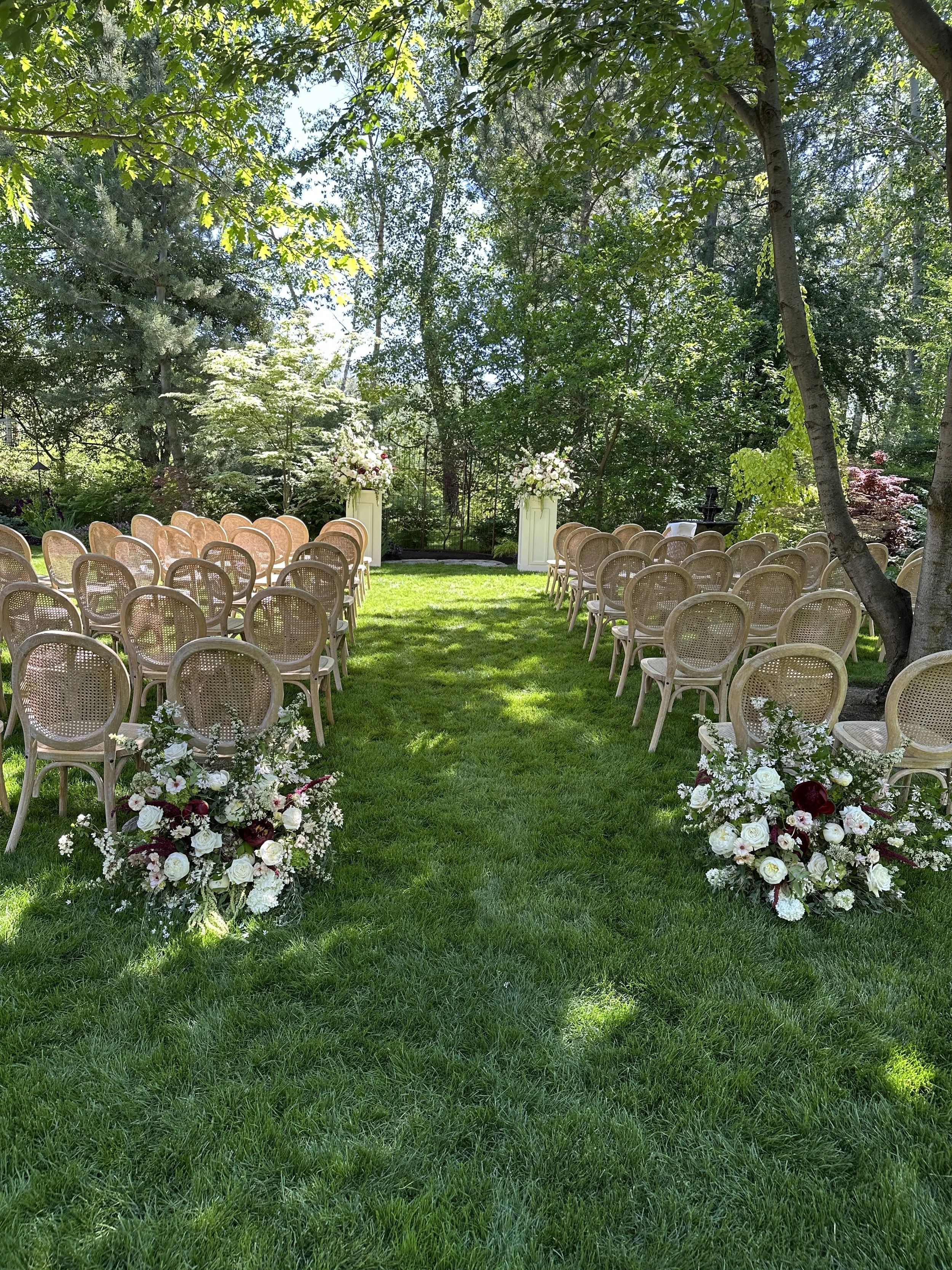 Outdoor wedding ceremony setup in a lush green garden with rows of wooden chairs facing an arch decorated with white and pink flowers, surrounded by trees and foliage. Created by Boise wedding florist at Edwards Flowershop.