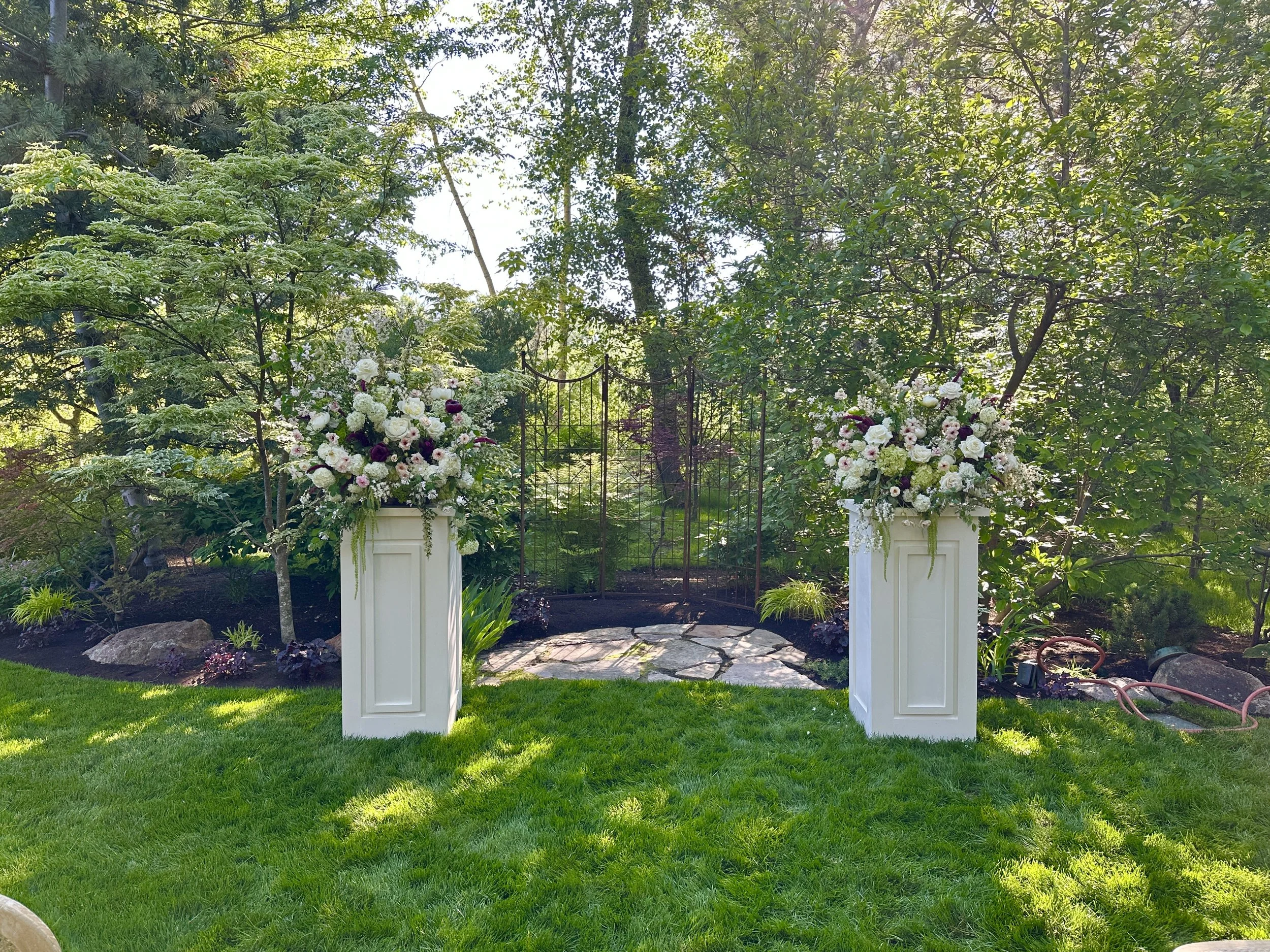 A garden wedding scene with two white pedestals holding floral arrangements of white and dark purple flowers, set on a stone pathway in front of a black wrought iron garden gate. Created by Boise wedding florist at Edwards Flowershop.