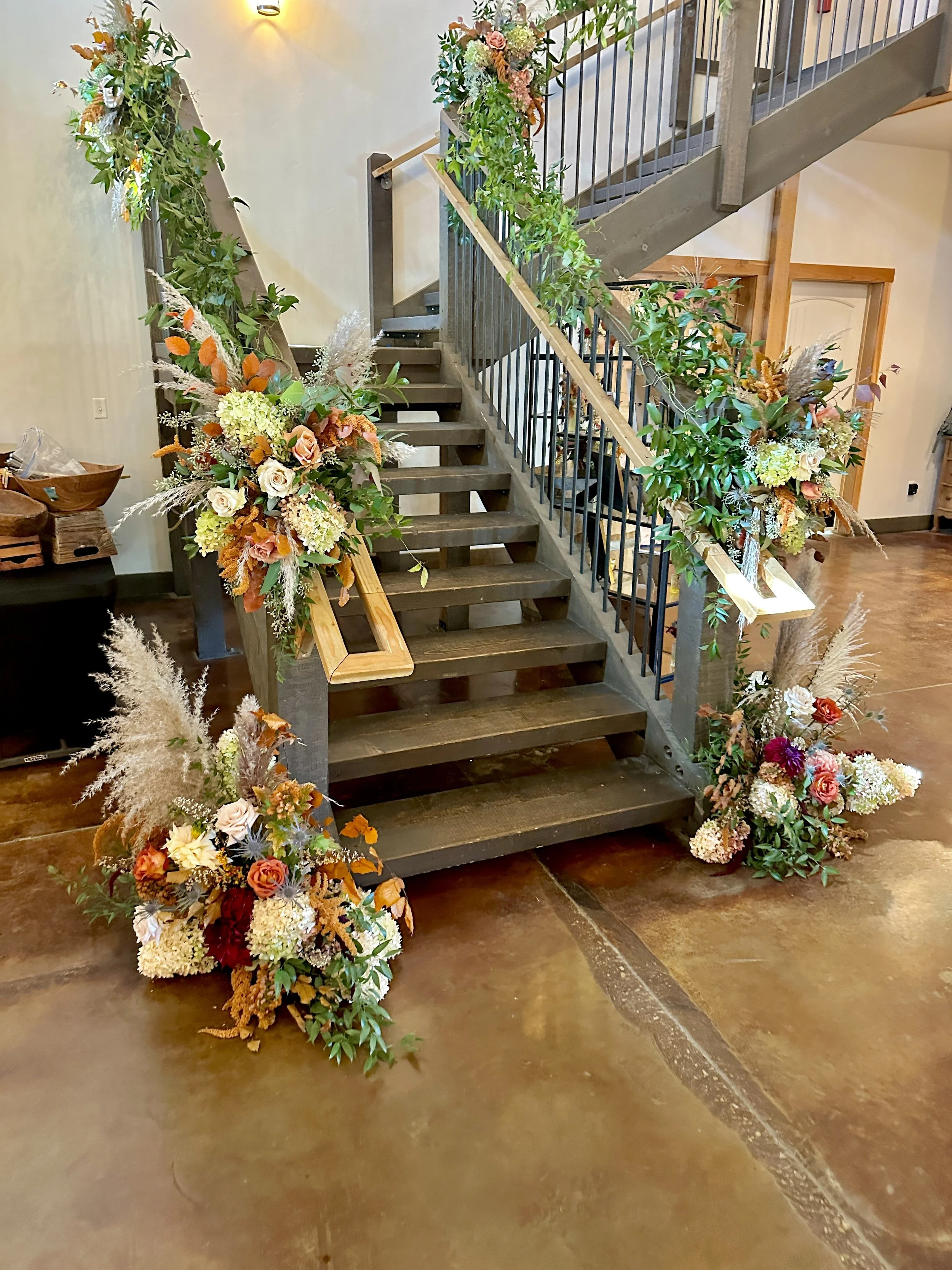 Decorated staircase with floral arrangements on the handrails and at the base, featuring various colorful flowers and greenery in a warm indoor setting, for a wedding reception. Wedding flowers by Boise florist Edwards Flowershop.