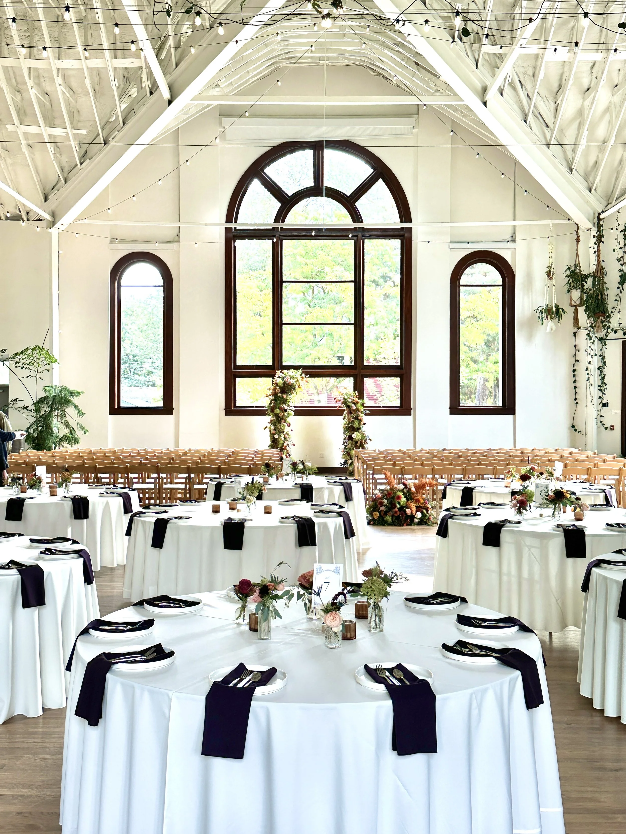 Wedding reception flowers featuring a large two piece arch backdrop and floral centerpieces on each table, created by Boise florist Edwards Flowershop. The room has large arched windows, wooden floors, and hanging string lights along the ceiling.