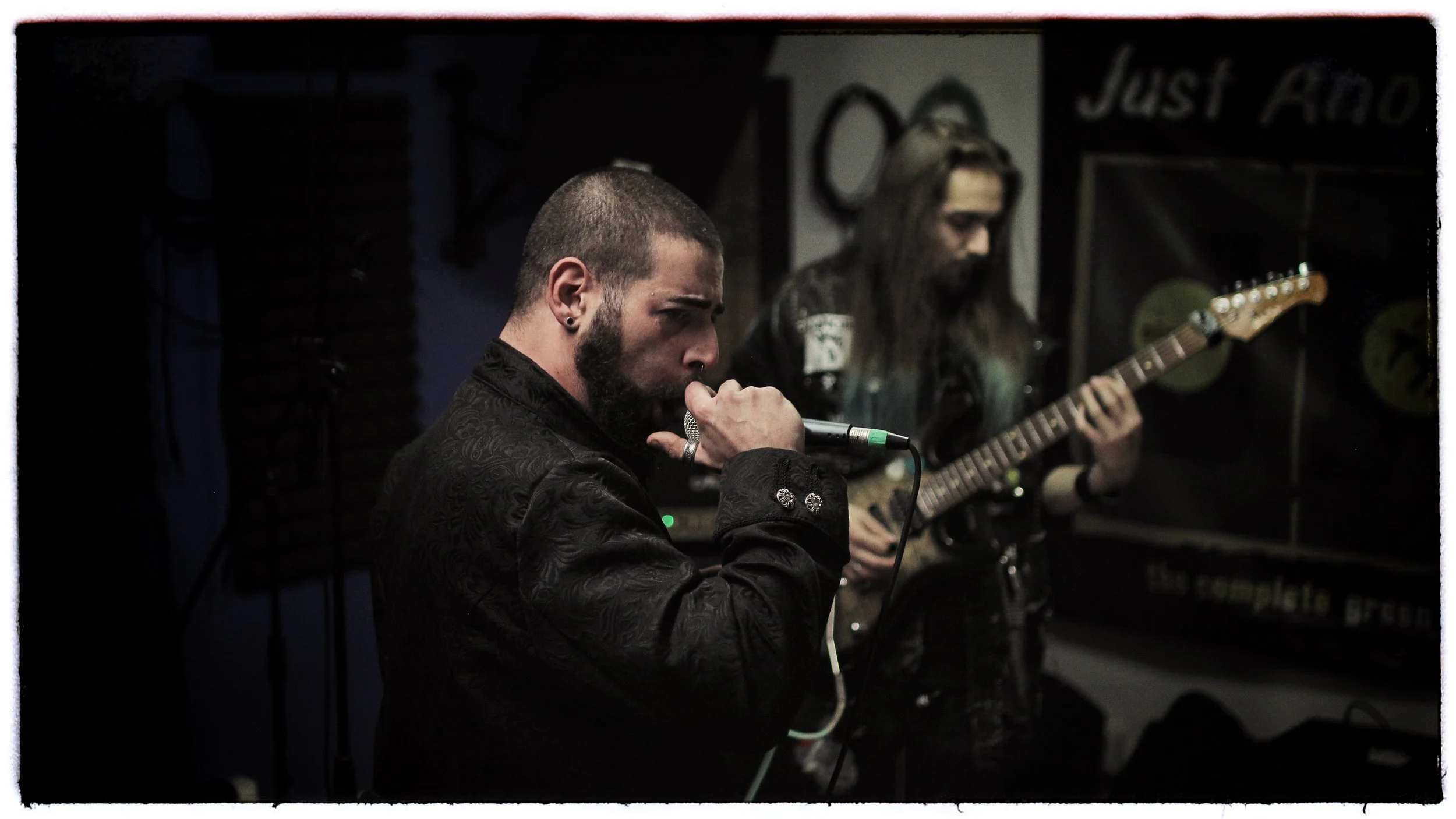 Musicians performing in a dimly lit venue. The foreground shows a man with short dark hair, a beard, wearing a patterned black jacket, holding a microphone close to his mouth. The background features a woman with long hair, playing an electric guitar