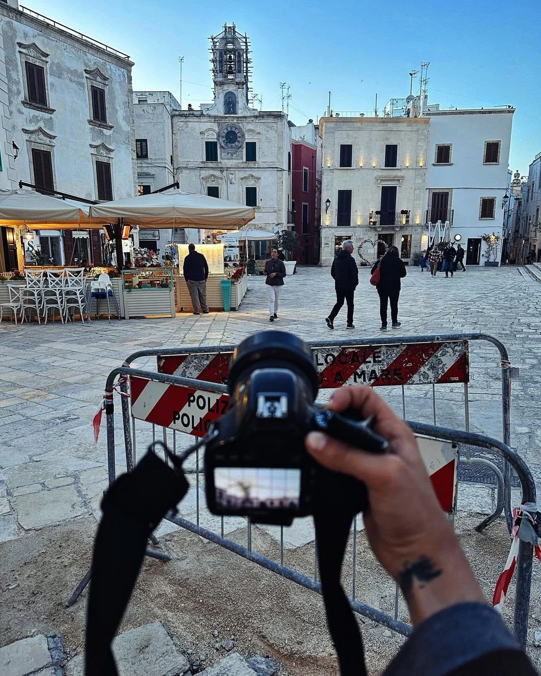 Person taking a photo of a town square with a camera, barricades with 'POLIZEI POLICE' sign, historic buildings, some people walking, outdoor cafe, in the background a church tower under a clear sky.