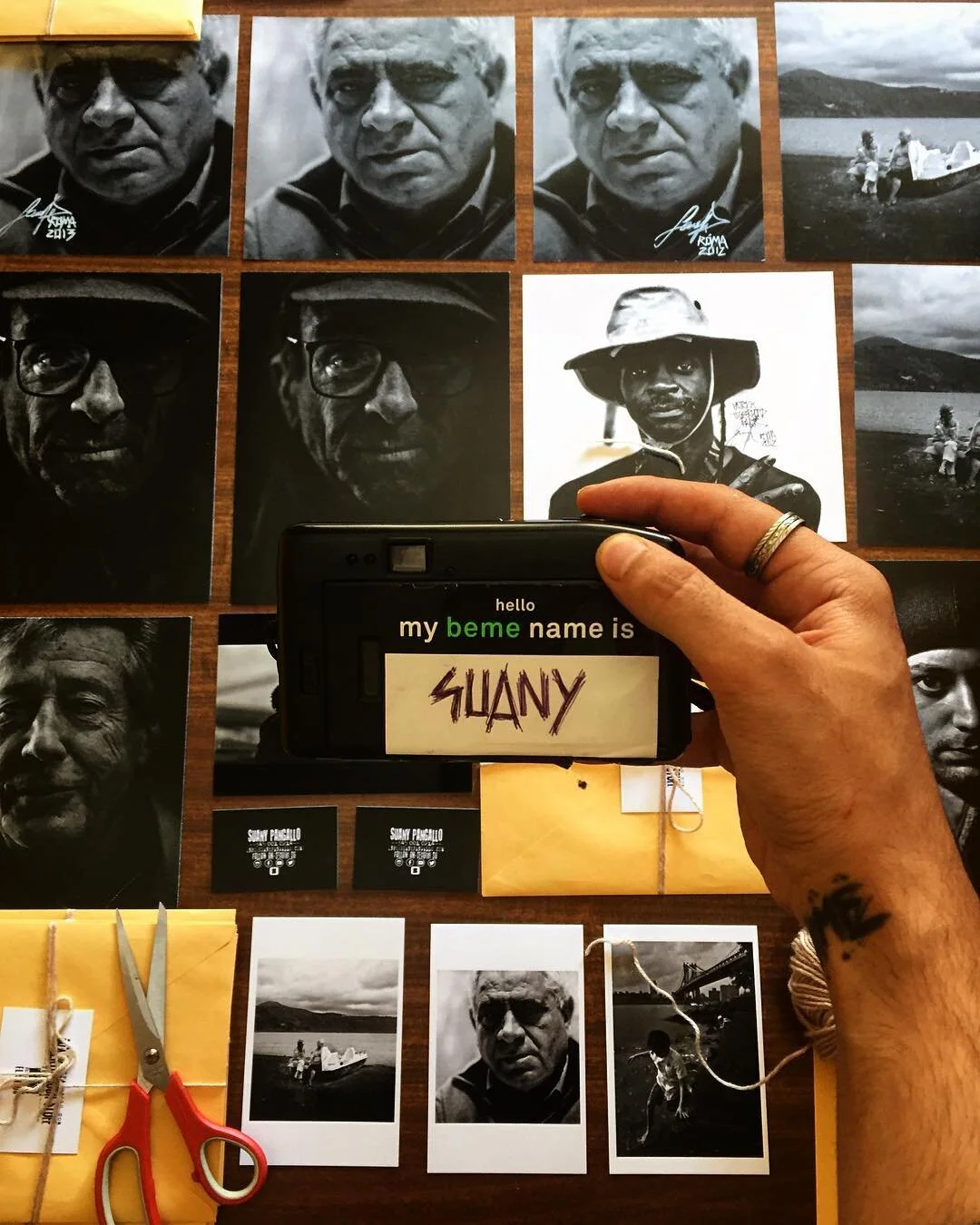 A person is holding a small electronic device displaying the name 'SUANY' on a digital screen, in front of a display of black-and-white portrait photographs of an older man, along with other photographs of a boat, a bridge, and people outdoors on a l