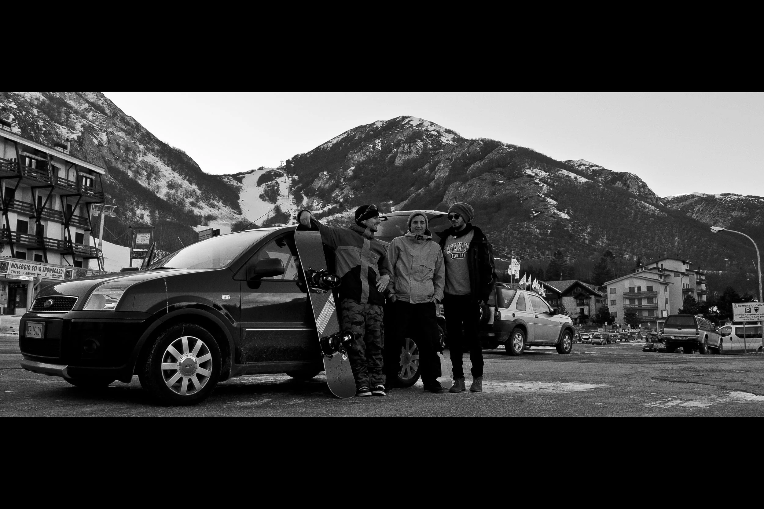Three men in winter clothing standing beside a parked car with a mountain landscape in the background.