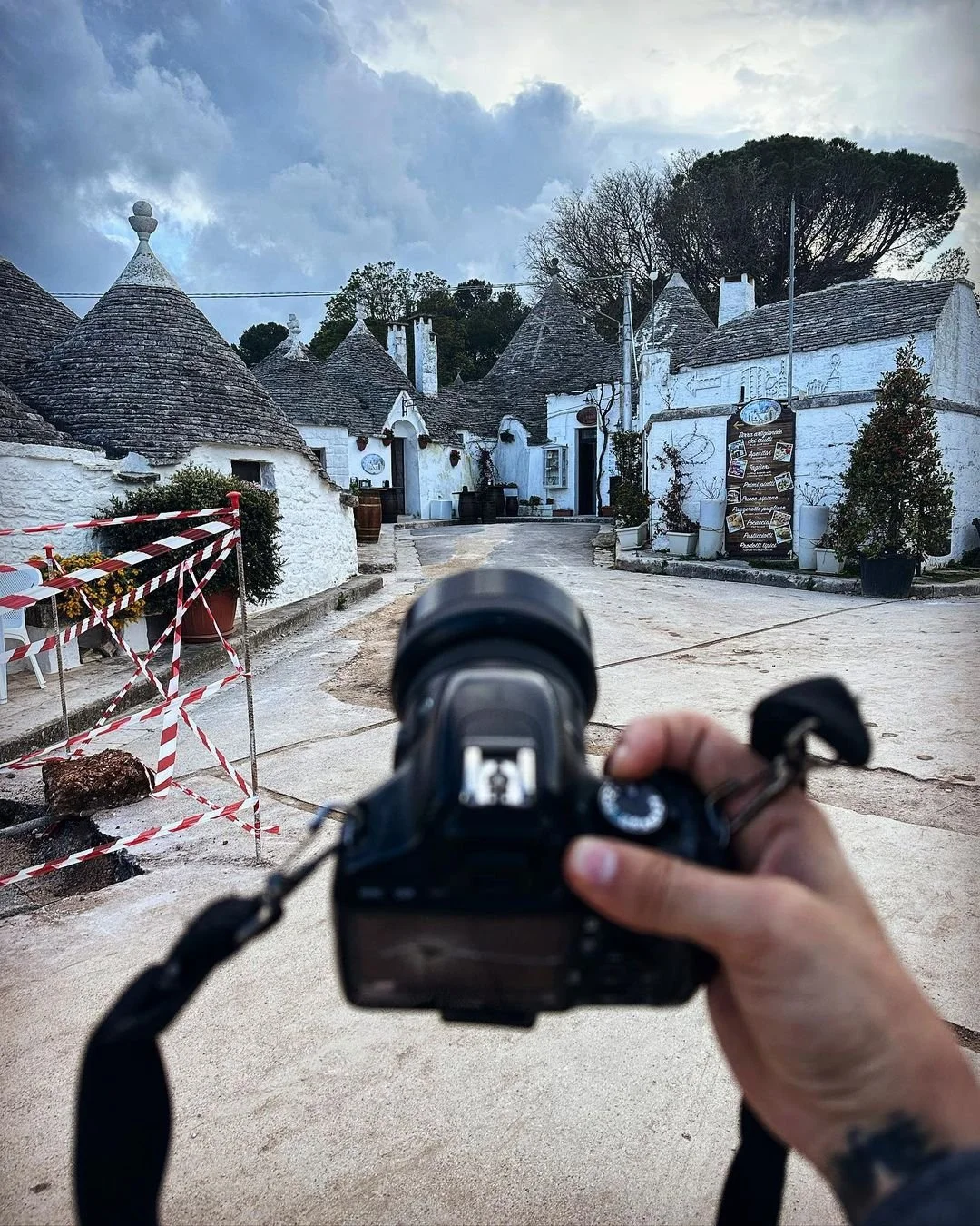 A person holding a camera in front of a picturesque village of white buildings with conical roofs, possibly in Italy, with cloudy sky overhead.