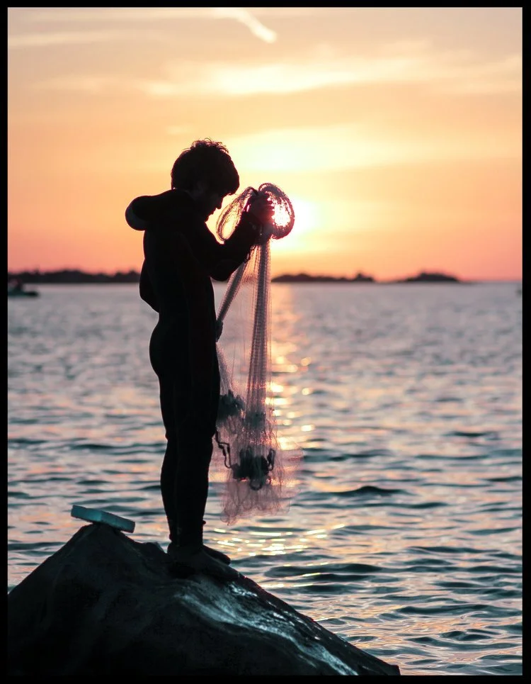 Silhouette of a person standing on a rock, fishing with a net at sunset over a body of water.