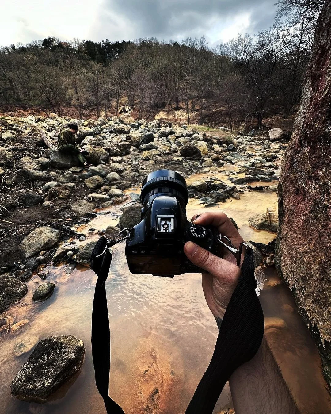 Photograph of a person holding a camera, aimed at a rocky creek with a person sitting on a rock in the background, surrounded by leafless trees and overcast sky.