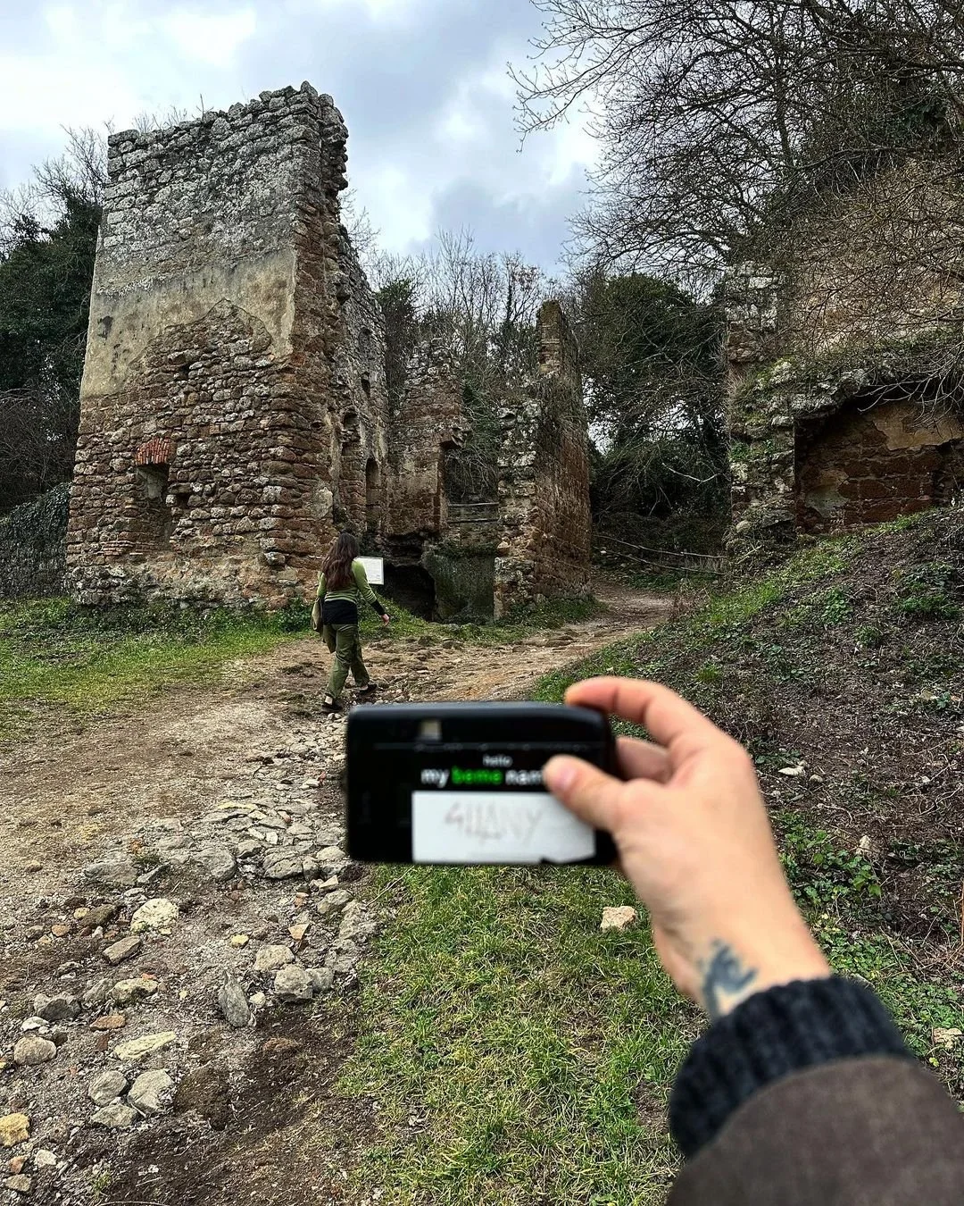 Person holding a smartphone in front of ruins made of stones and bricks, with another person walking on a stone path toward the ruins, surrounded by trees and a cloudy sky.