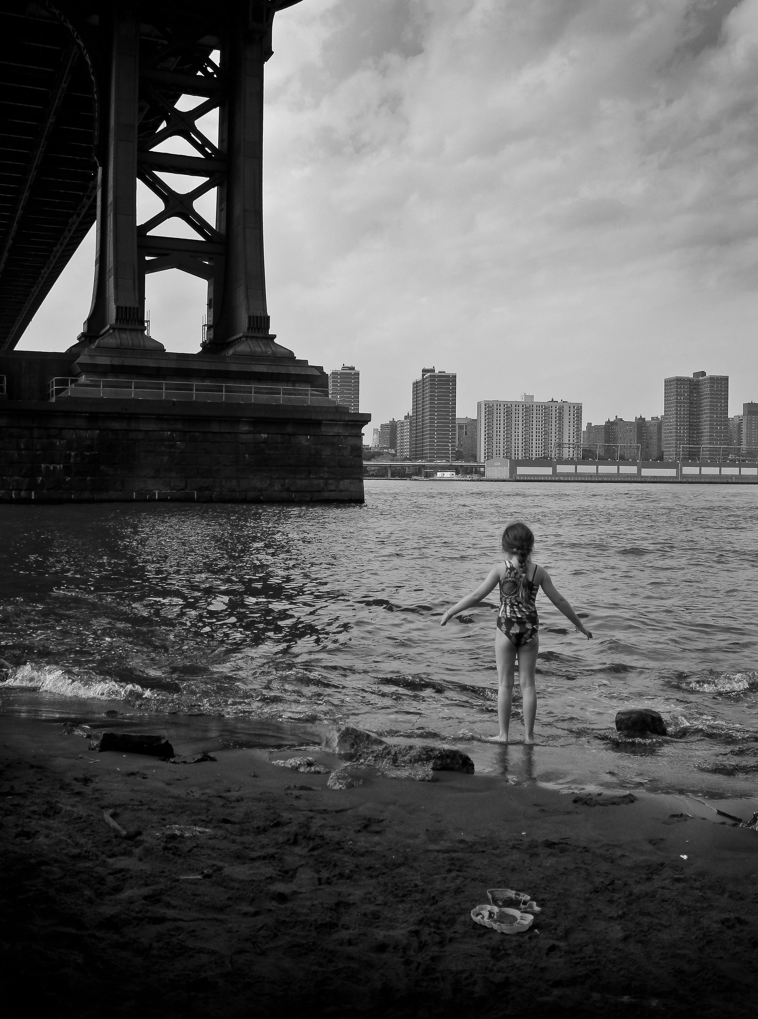 A girl in a swimsuit plays near the water on a sandy riverbank under a bridge, with a city skyline of tall buildings in the background. The photo is in black and white.