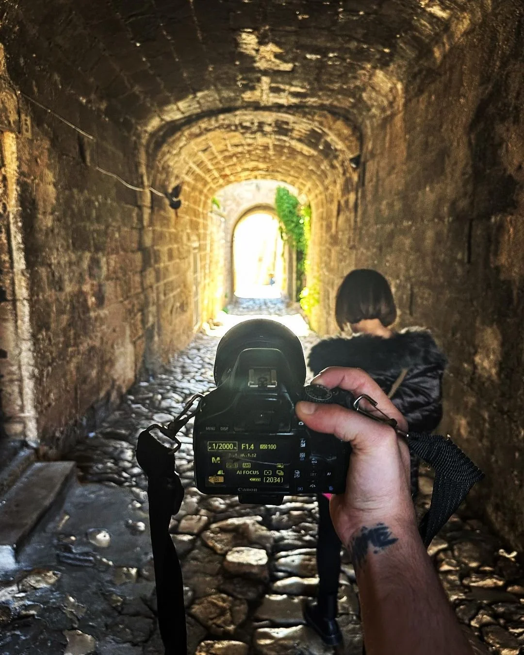 Looking through a camera at a narrow stone tunnel with cobblestone floor and sunlight at the end.
