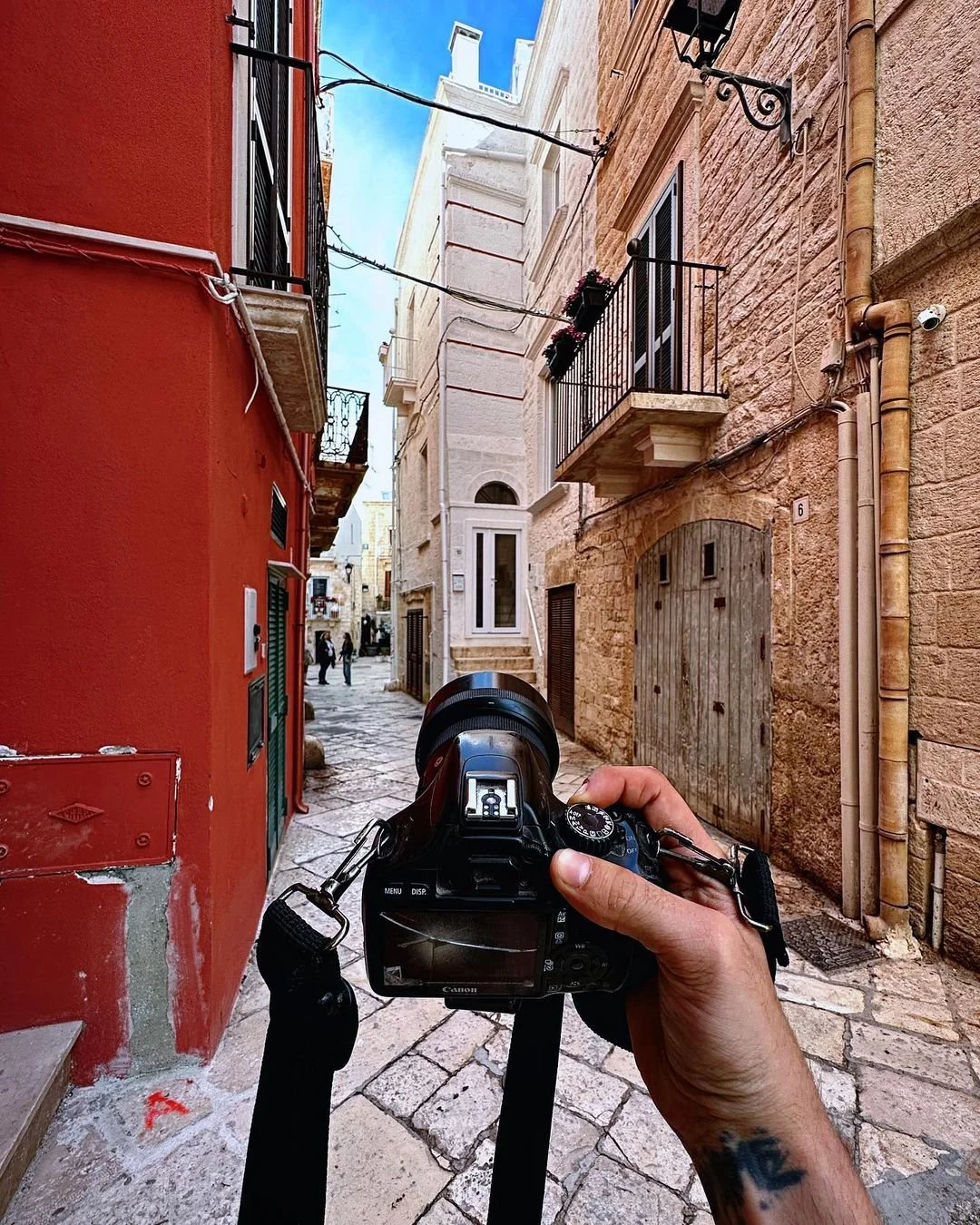 A person holding a Canon DSLR camera captures a narrow, cobblestone street with colorful buildings, black wrought iron balconies, and potted plants in a European town.