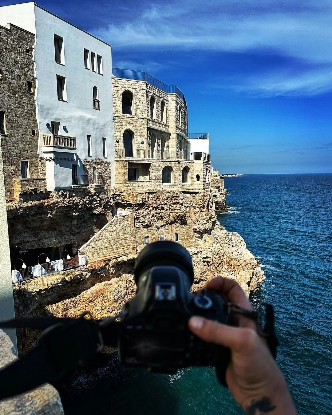 A hand holding a camera takes a photo of buildings on a rocky coastline with the ocean in the background.