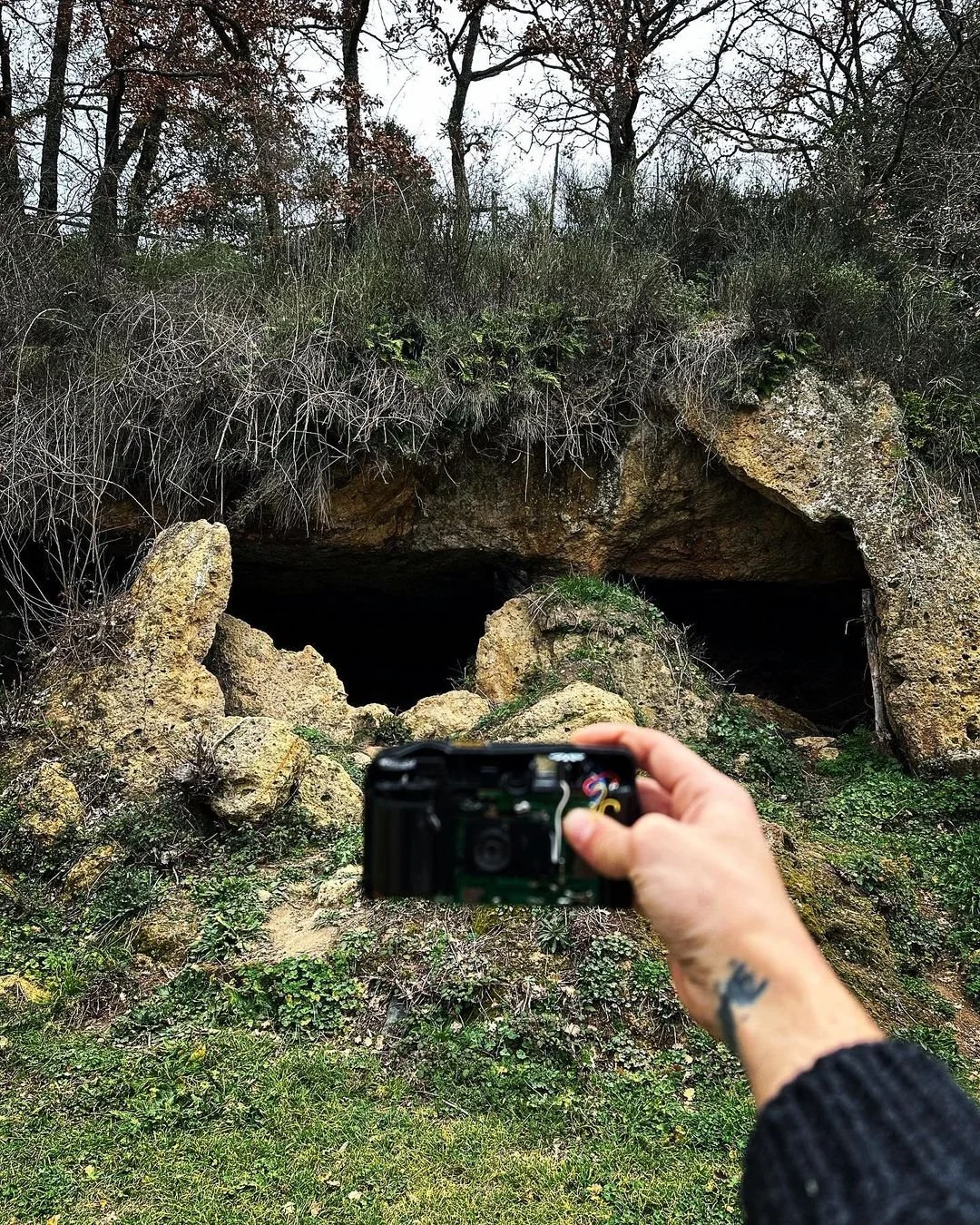Person holding a camera in front of a rocky cave entrance surrounded by vegetation and trees.