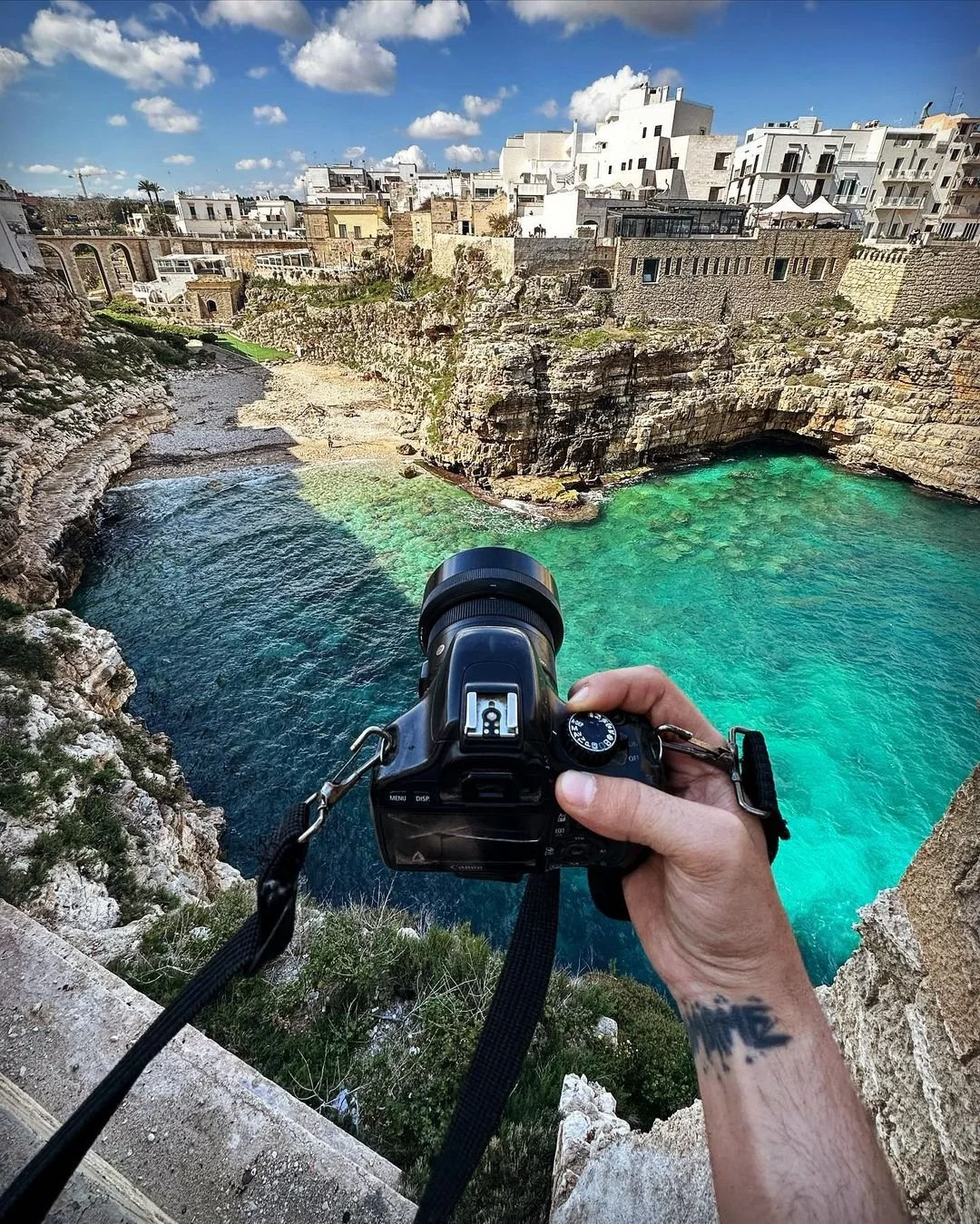 A person holding a camera takes a photo of turquoise water, rocky cliffs, and white buildings under a partly cloudy sky.