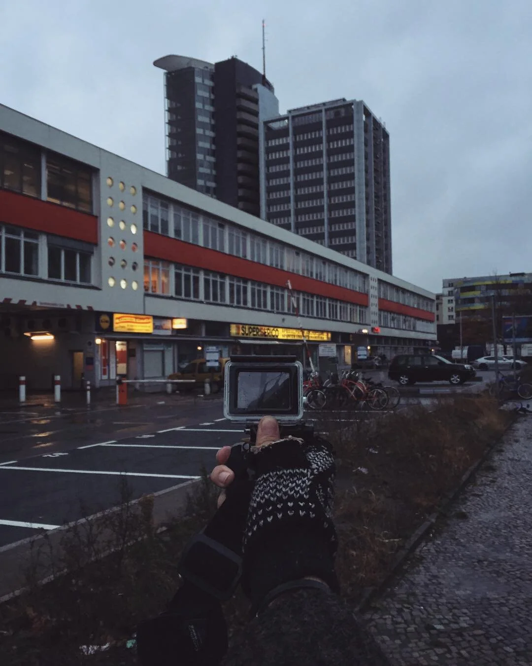 A person wearing black gloves with white patterns is holding a camera or GoPro, capturing the scene of tall buildings in a city during dusk, with parked cars and a wet street in the background.