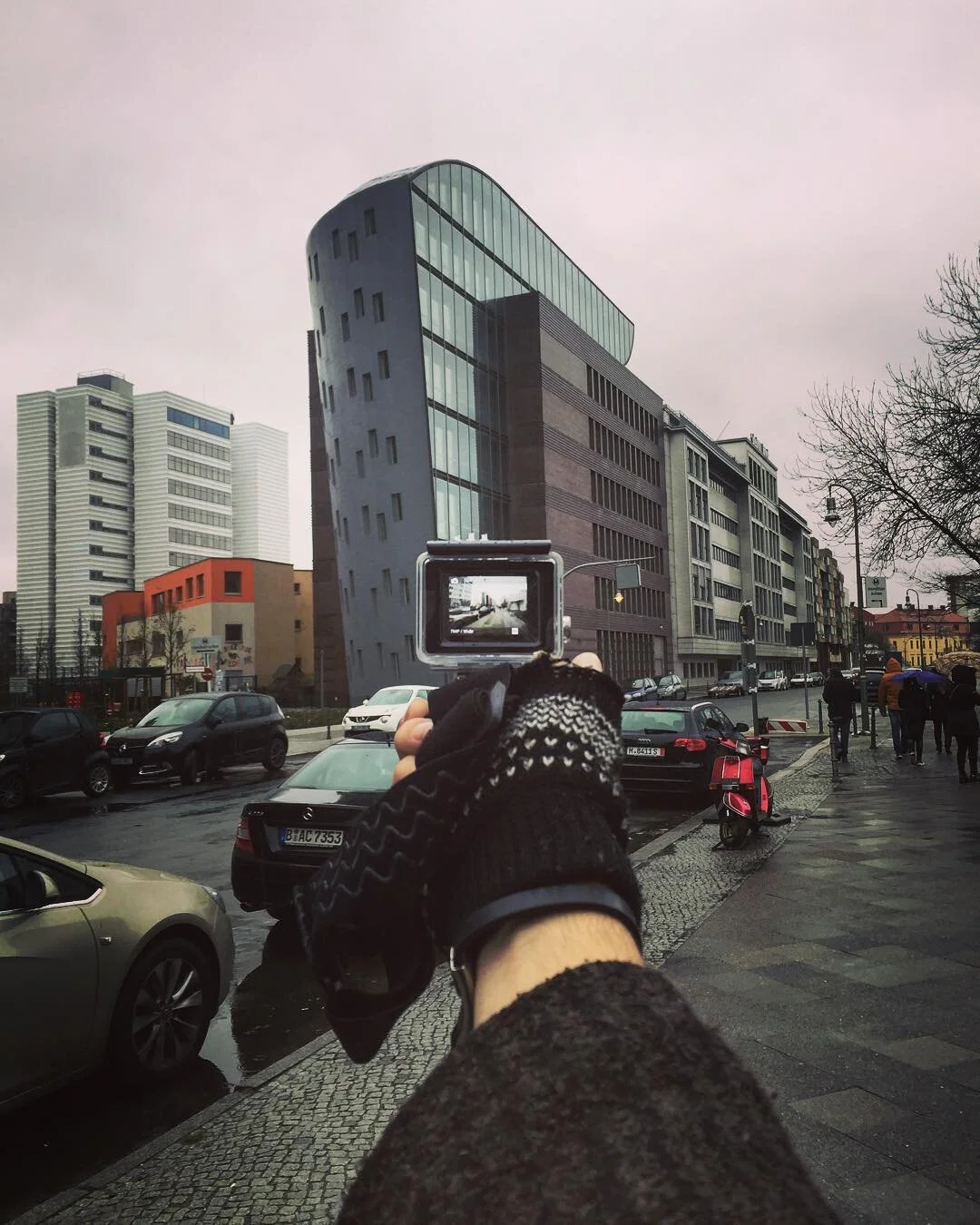 Hand holding a camera taking a photo of a modern, uniquely shaped building in an urban street scene with cars and pedestrians on a cloudy day.