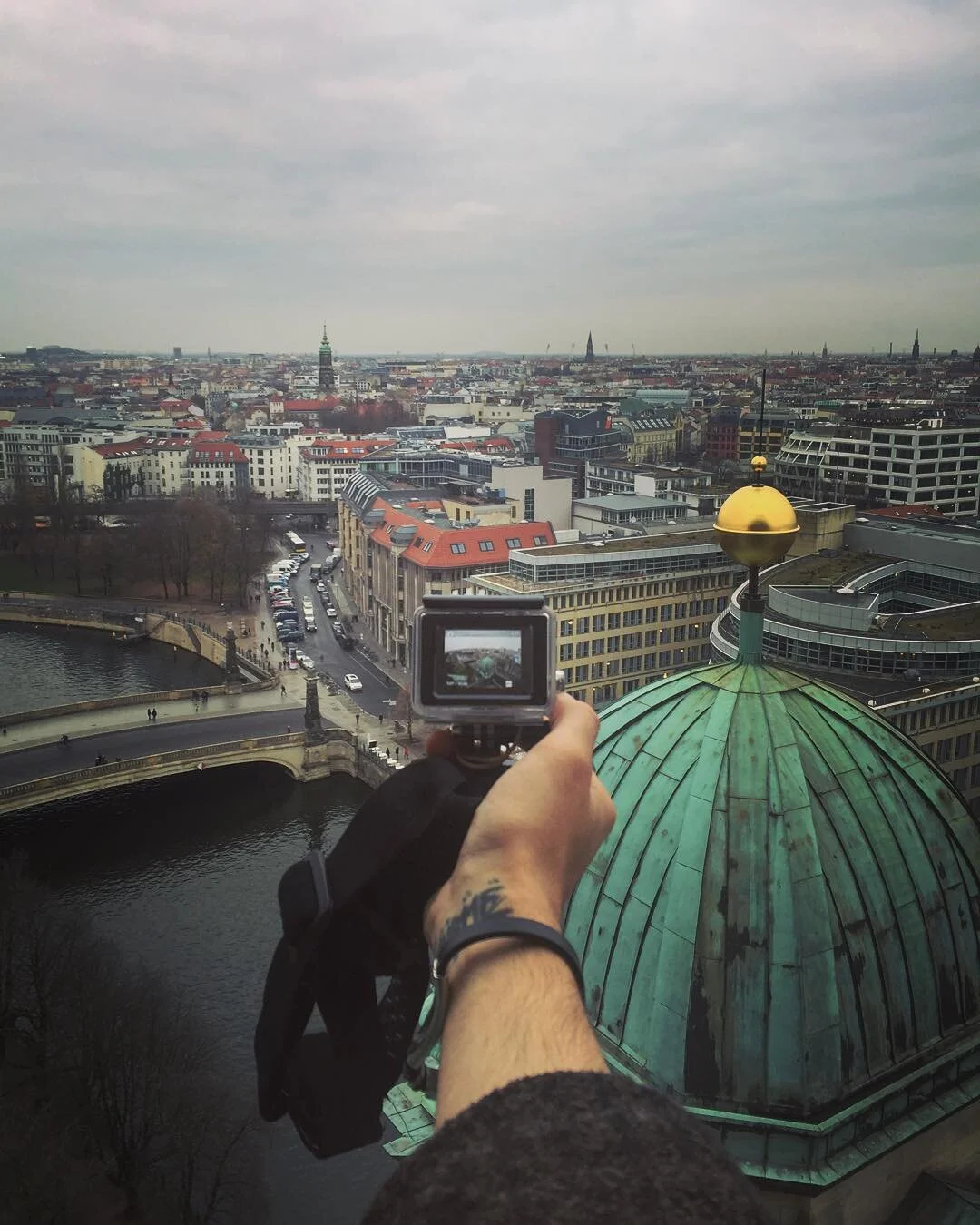 Person taking a photo of a cityscape from a high vantage point, showing rooftops, a river, and buildings in a city under an overcast sky.