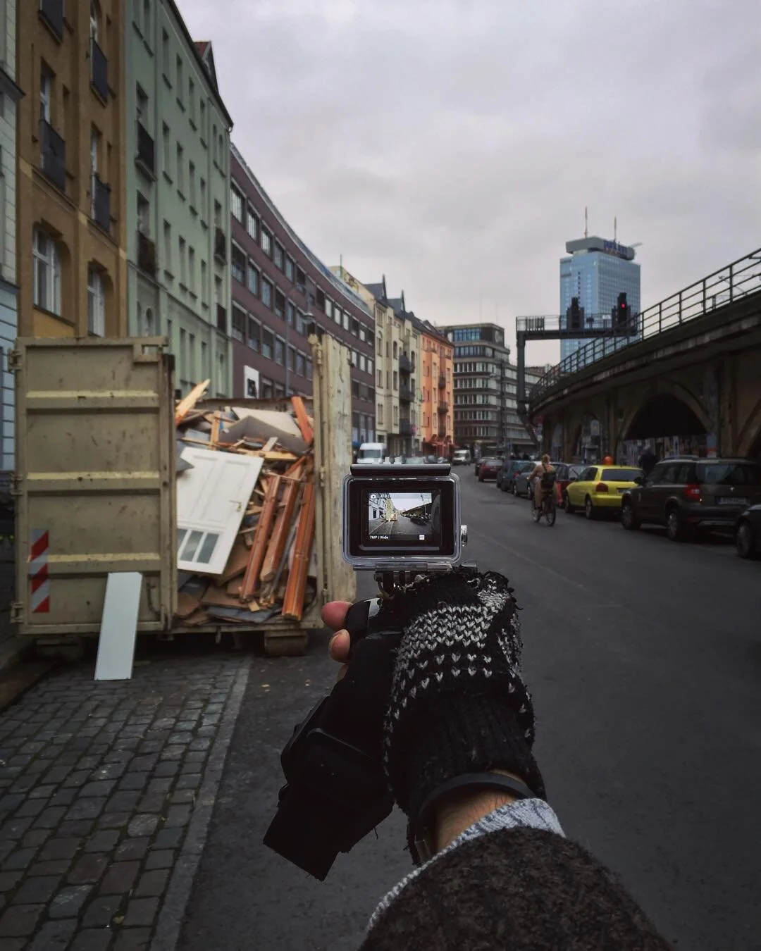 Person wearing black and white glove holding a camera, capturing a city street scene with parked cars, colorful buildings, and a tall blue skyscraper in the background.