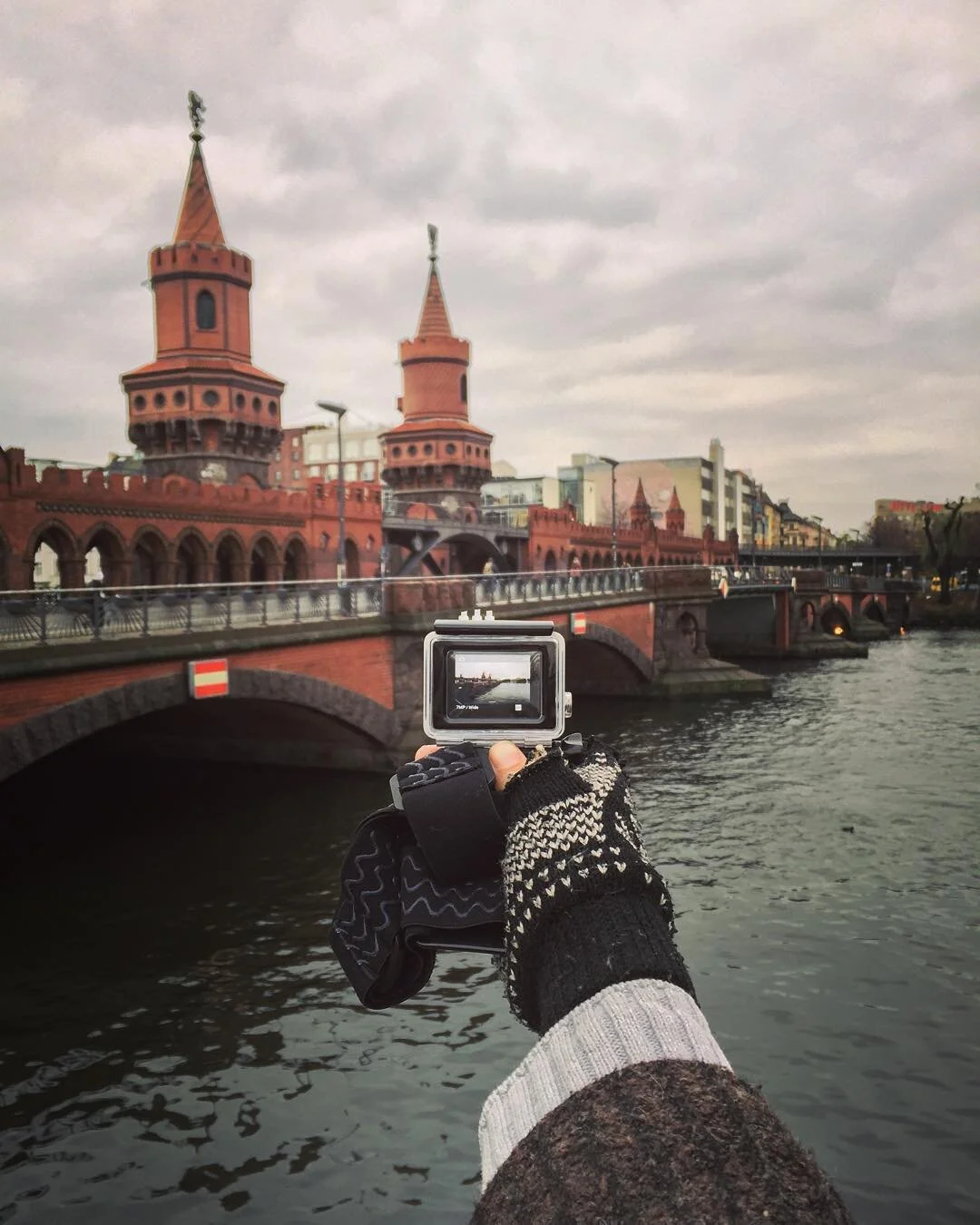 A person wearing black and white patterned gloves and a gray sweater is taking a photo with a camera. The photo shows a historic red brick bridge with twin towers over a body of water, under a cloudy sky.