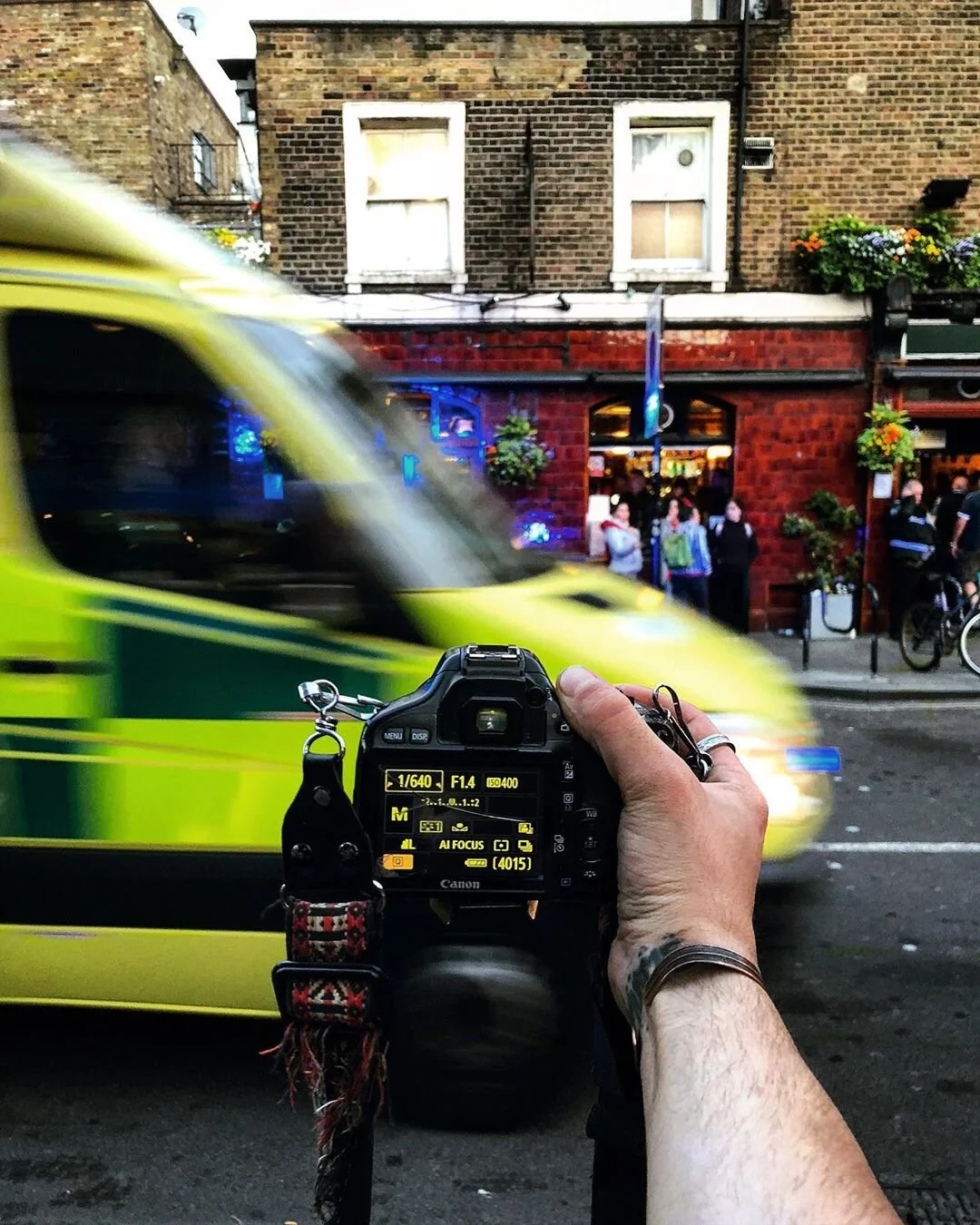 A person holding a Canon DSLR camera is capturing a photo of a moving yellow and green emergency vehicle passing by on a city street. The background shows a red brick building with a pub entrance, people walking and standing outside, and hanging flow