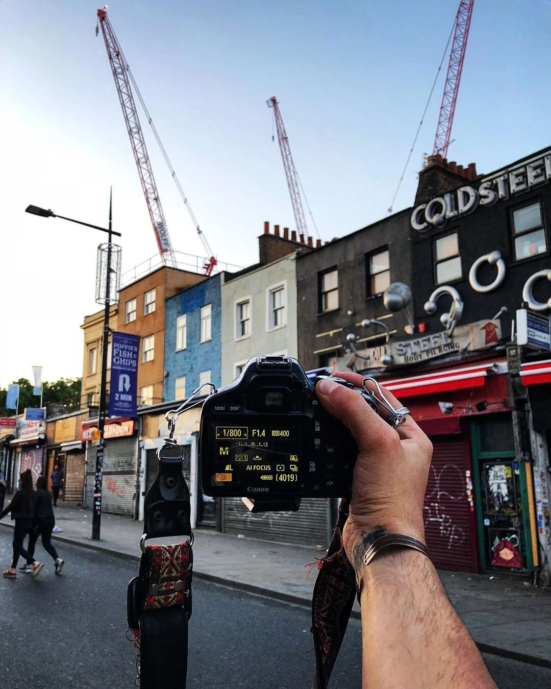 A person holding a camera taking a photo of a city street with colorful buildings, cranes, and a sign that reads Cold Steel.