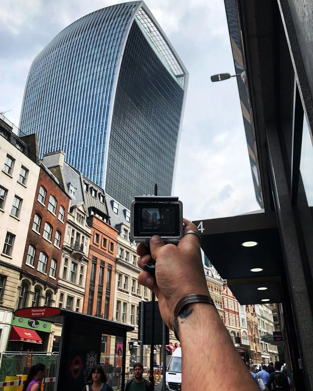 Person taking a photo of a tall modern skyscraper with a curved glass exterior in a city street, with older buildings and pedestrians below.
