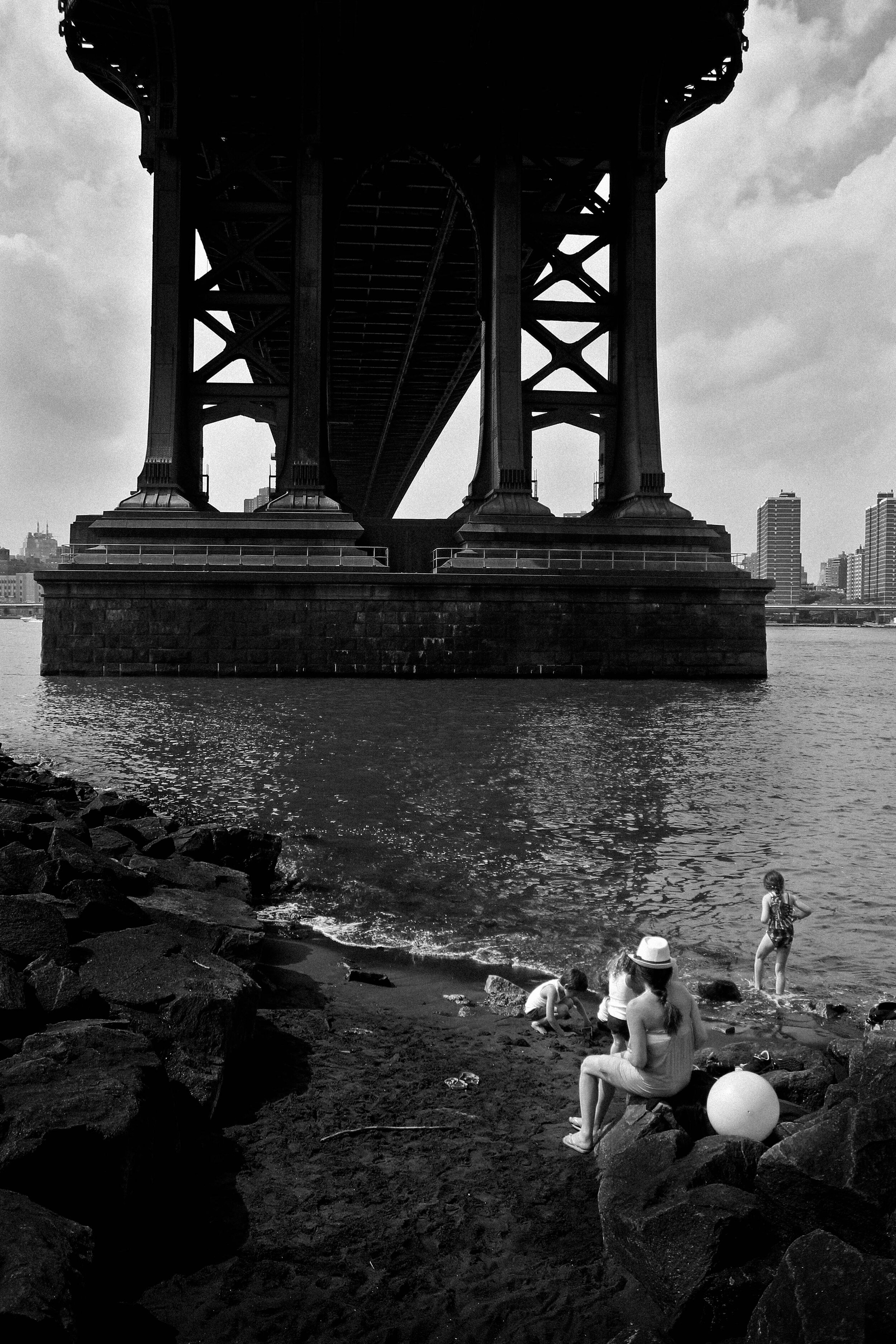 People sitting and playing near the water on a rocky shoreline under a bridge in an urban setting.