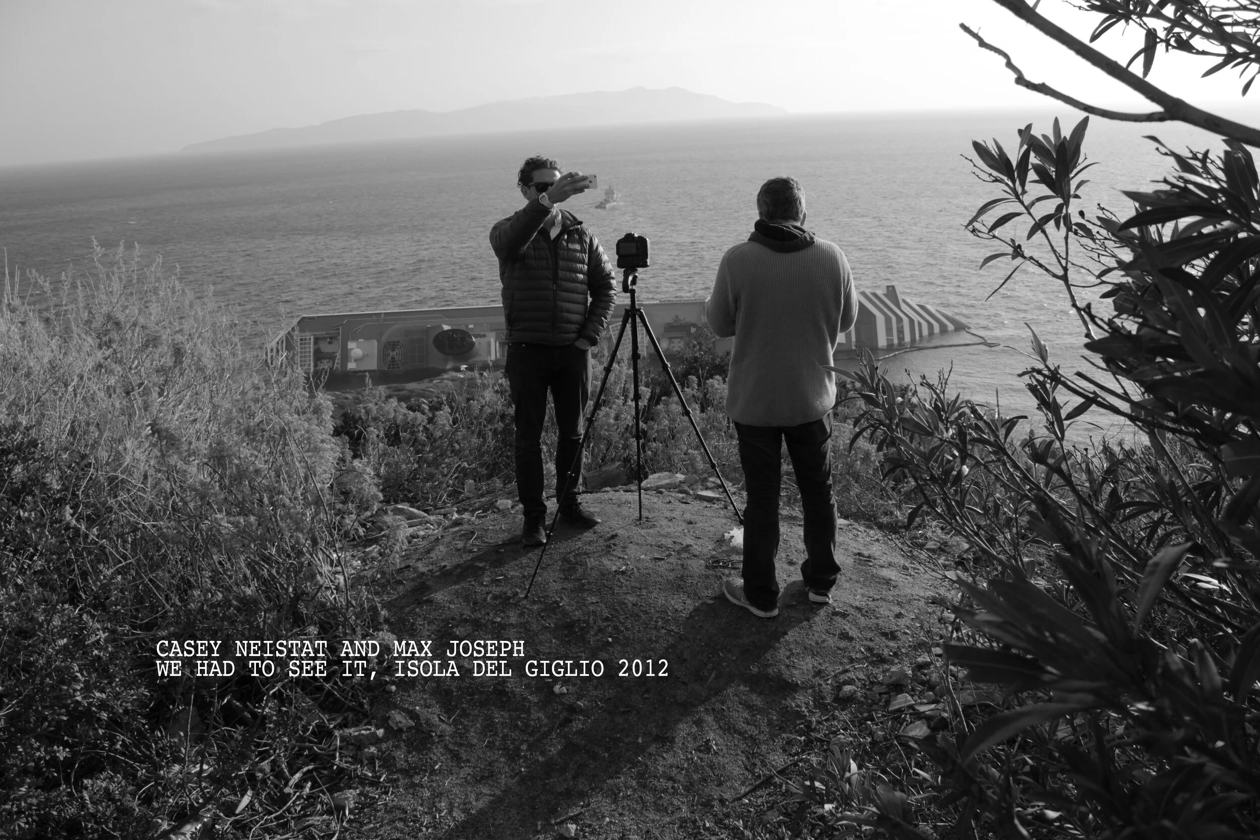 Two men with a camera set up on a tripod, one taking a photo of the ocean view with an island in the distance, framed by bushes and trees in a coastal setting, black and white photo, with an overlay caption about Cassy Netstat and Max Joseph at Isola del Giglio in 2012.