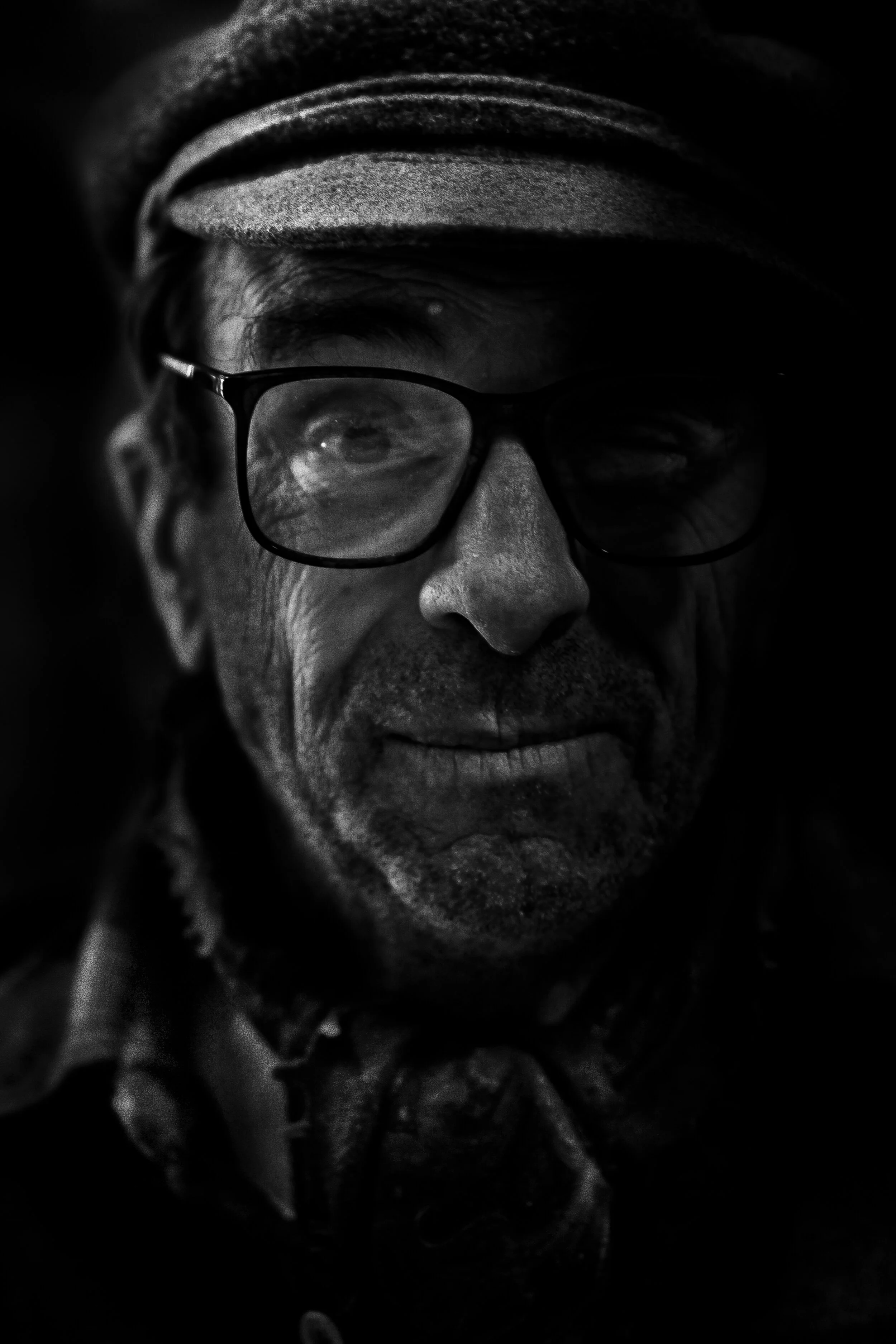 Close-up black and white photo of a person wearing glasses, with a hat and a contemplative expression, in a dark setting.