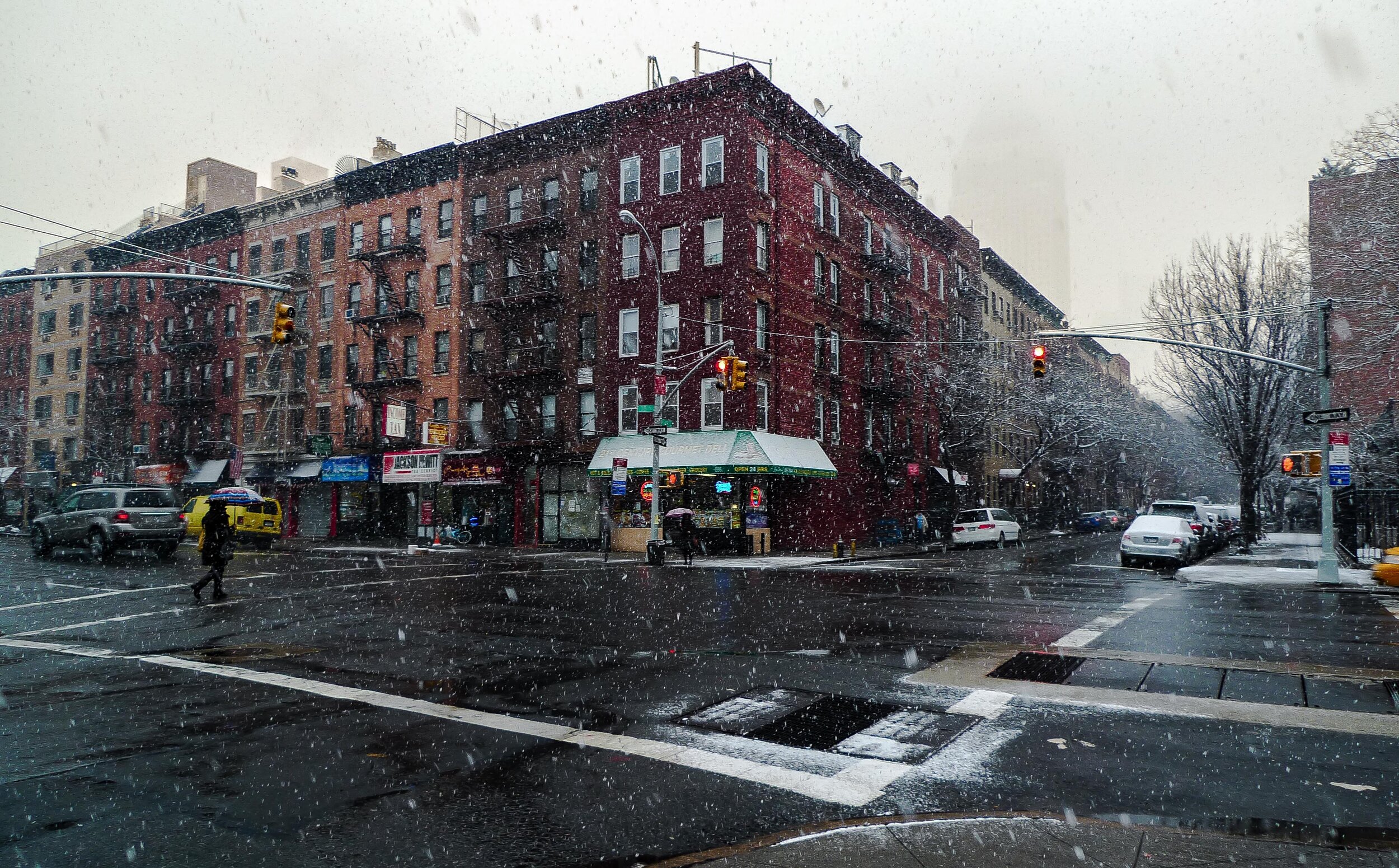 A city intersection during snowfall with cars and pedestrians. Multi-story brick buildings line the street, and traffic lights show red. Some pedestrians have umbrellas.