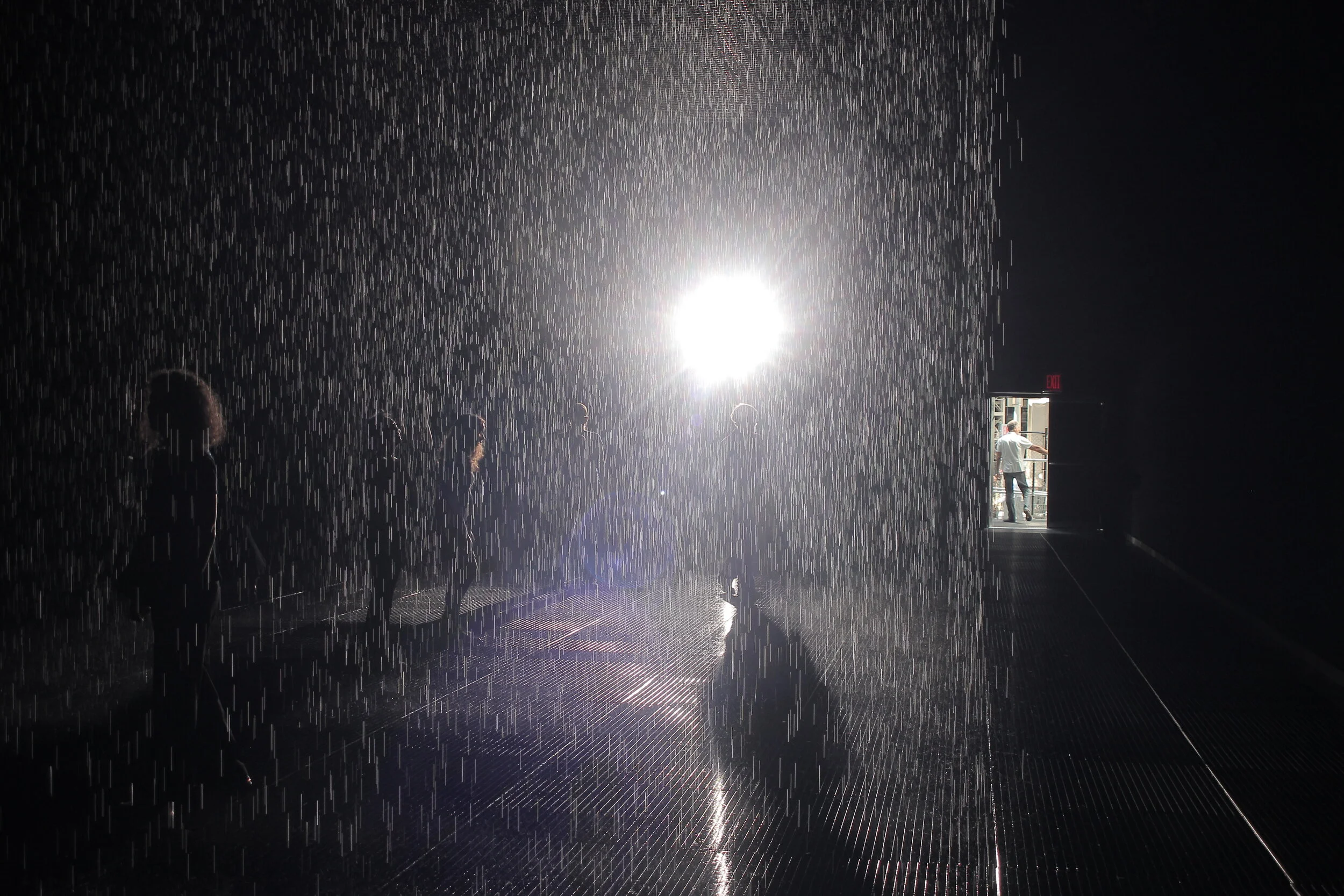People standing under a rain of water illuminated by bright lights in a dark indoor space, with a doorway showing outside.