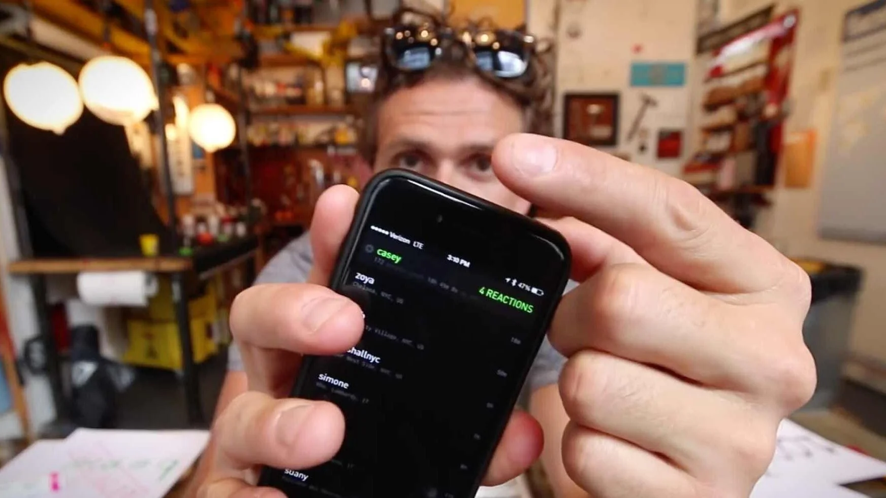 Person holding a smartphone in front of their face, with a cluttered bookshelf and warm lighting in the background.