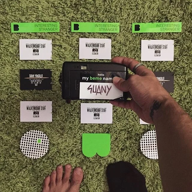 Arrangement of various small cards and objects on a green carpet, including a camera with a word on it, and paper cutouts creating a face-like composition.