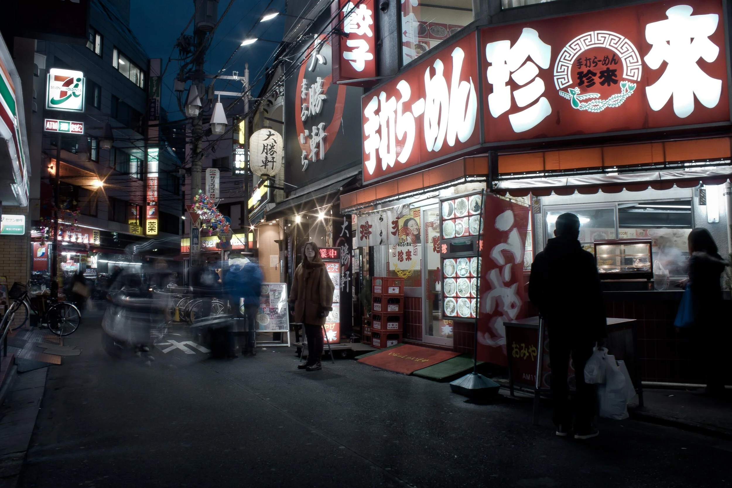 A busy city street scene at night with illuminated signs and storefronts in Japanese, including a ramen restaurant and a 7-Eleven. People are walking, and there is a steam pot visible at the ramen shop. Bicycles are parked on the side.