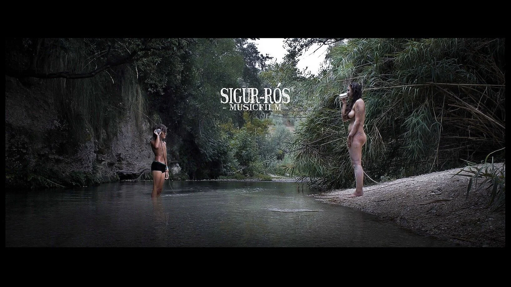 Two topless women, one standing in a river and the other on the riverbank, both drinking from bottles in a natural, wooded setting.