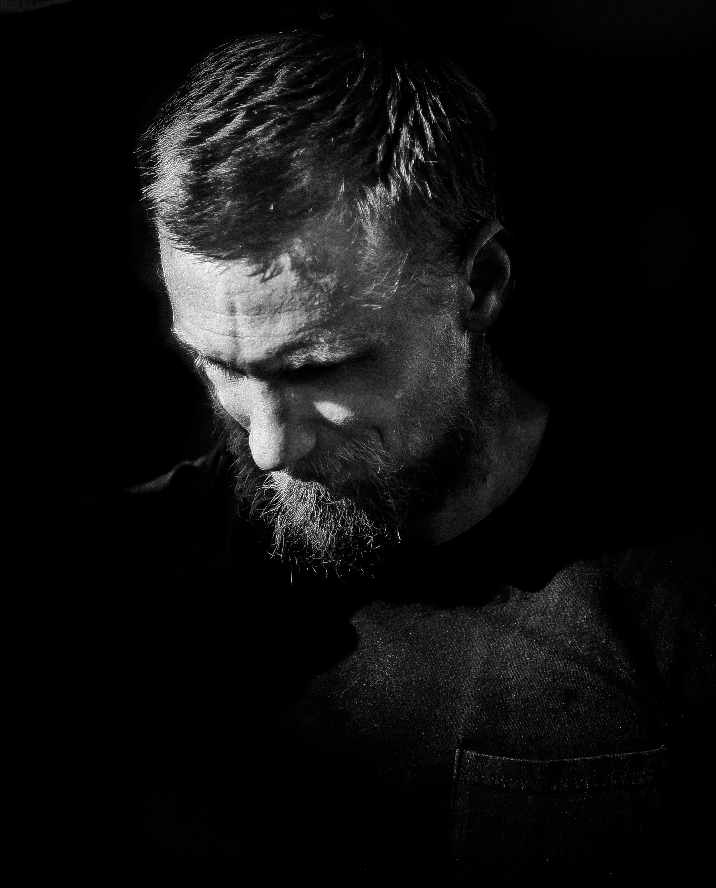 Black and white close-up of a man's face, looking downward with a beard and short hair, on a dark background.