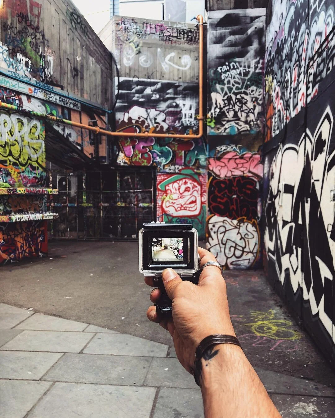 A person's hand holding a camera capturing a colorful graffiti-covered alley with various tags and murals on the walls.