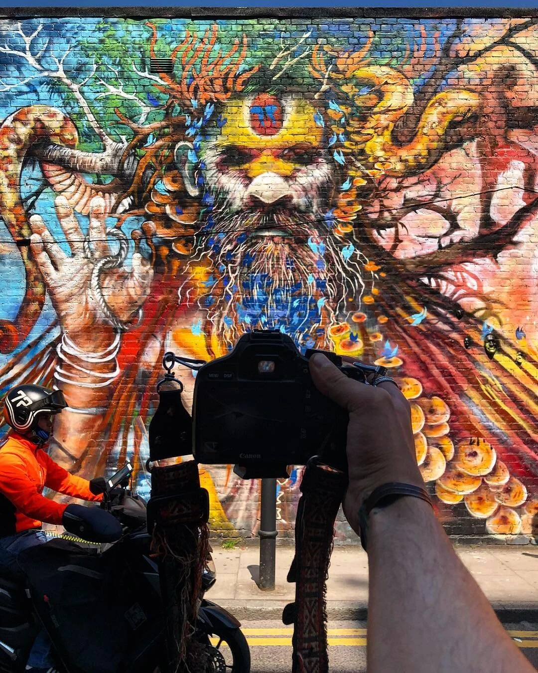 Colorful mural of a wise, bearded man with a third eye on his forehead, surrounded by branches, snakes, and vibrant abstract elements, with a person taking a photo and a motorcycle rider passing by in the foreground.