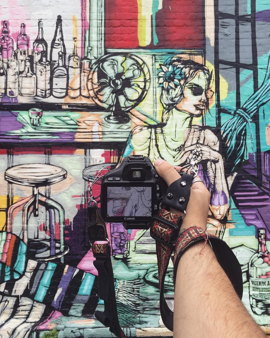 Close-up of a person holding a camera in front of colorful street art depicting a woman with flowers in her hair, sunglasses, and seated at a bar with bottles and a fan in the background.
