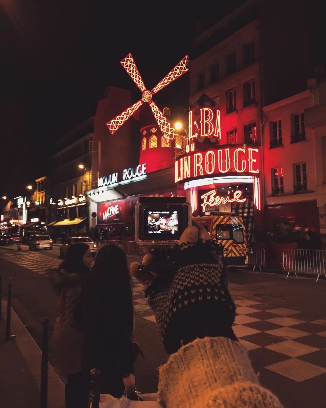 Nighttime scene on a city street with the neon-lit Moulin Rouge cabaret in the background, featuring a large windmill and red neon signs, with people taking photos in front of it.