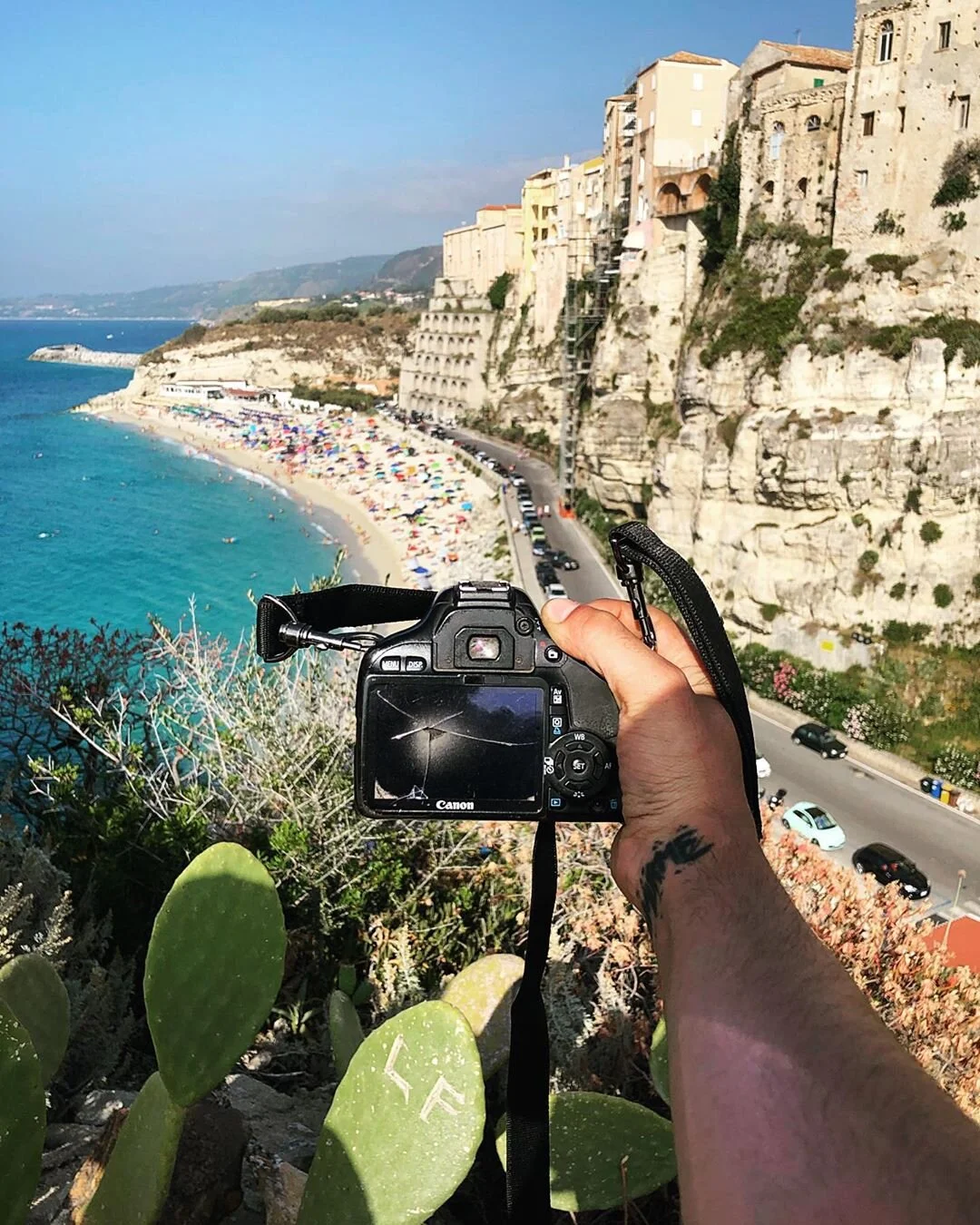 A person holding a Canon camera taking a photo of a beach with colorful umbrellas and cliffs with buildings above, along a coastal road.