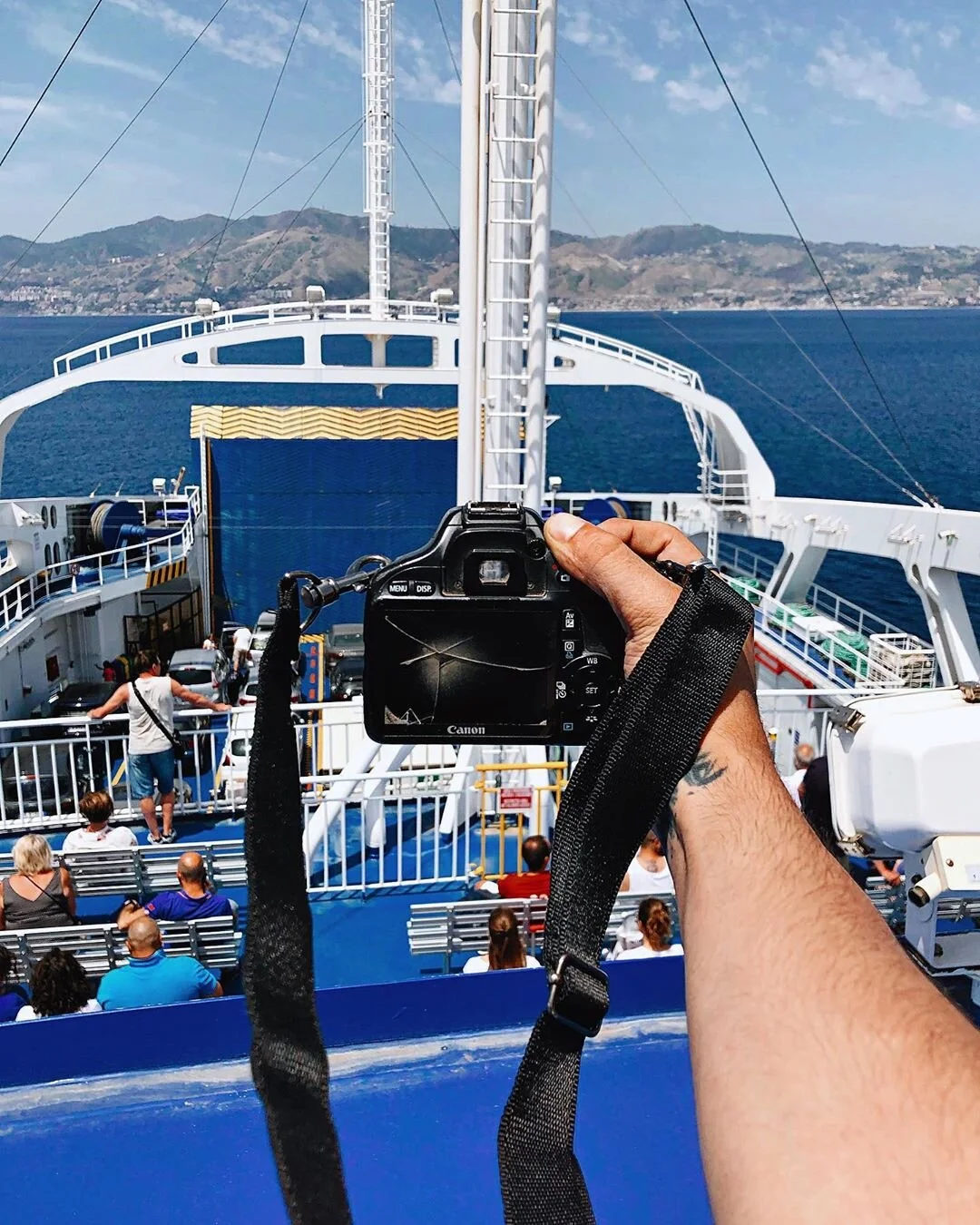 Person holding a camera taking a photo of the deck of a ferry boat with passengers on benches and people walking, ocean and distant mountains in the background.