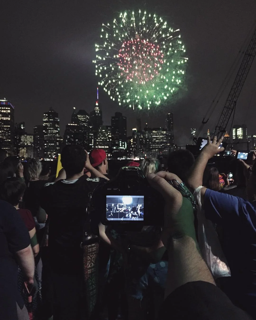 People watching fireworks display over a city skyline at night, with some taking photos.