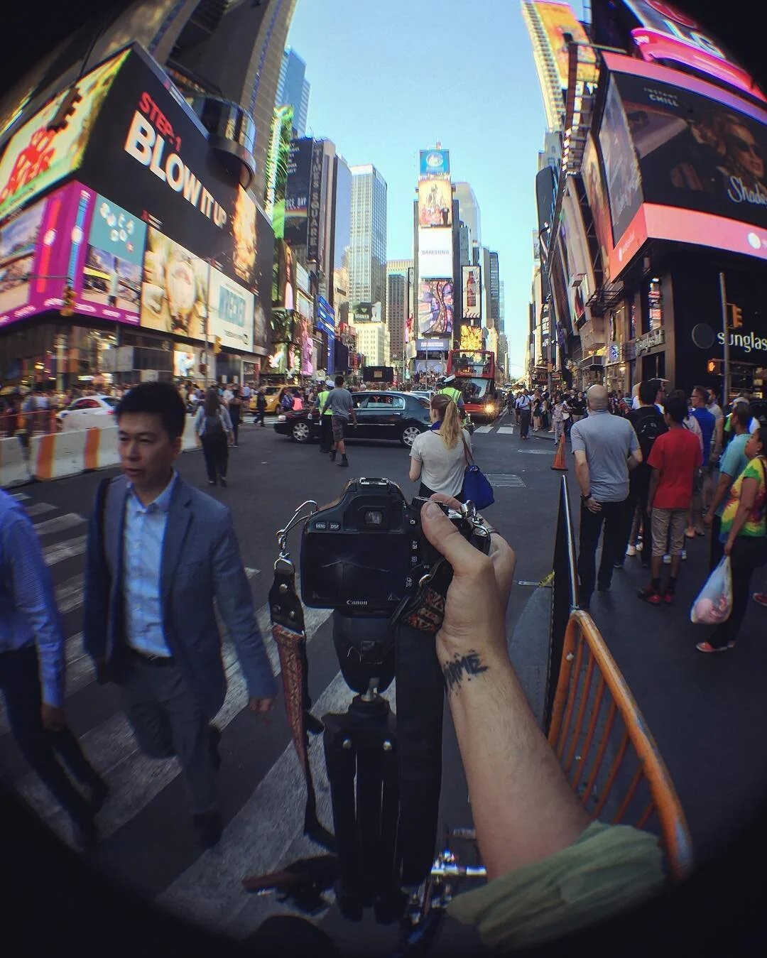Bright city street scene in Times Square with tall billboards, digital ads, and a crowd of pedestrians. The photo is taken from a person's perspective holding a camera, with their tattooed arm visible in the foreground.