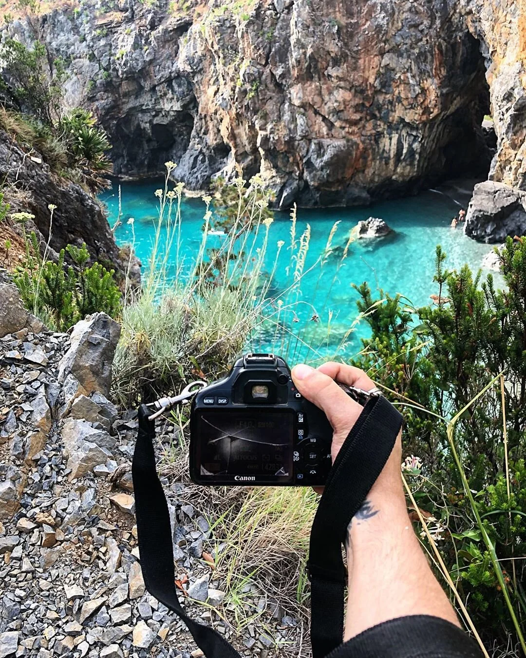 A person holding a camera is taking a photo of a turquoise water body surrounded by rocky cliffs and greenery.