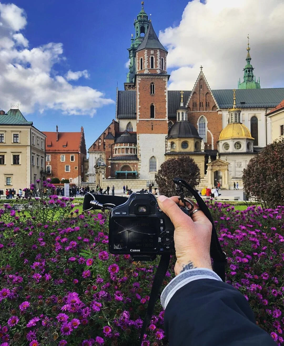A person taking a photo of a historic church or cathedral with a Canon camera, surrounded by purple flowers in a landscaped area, under a partly cloudy blue sky.