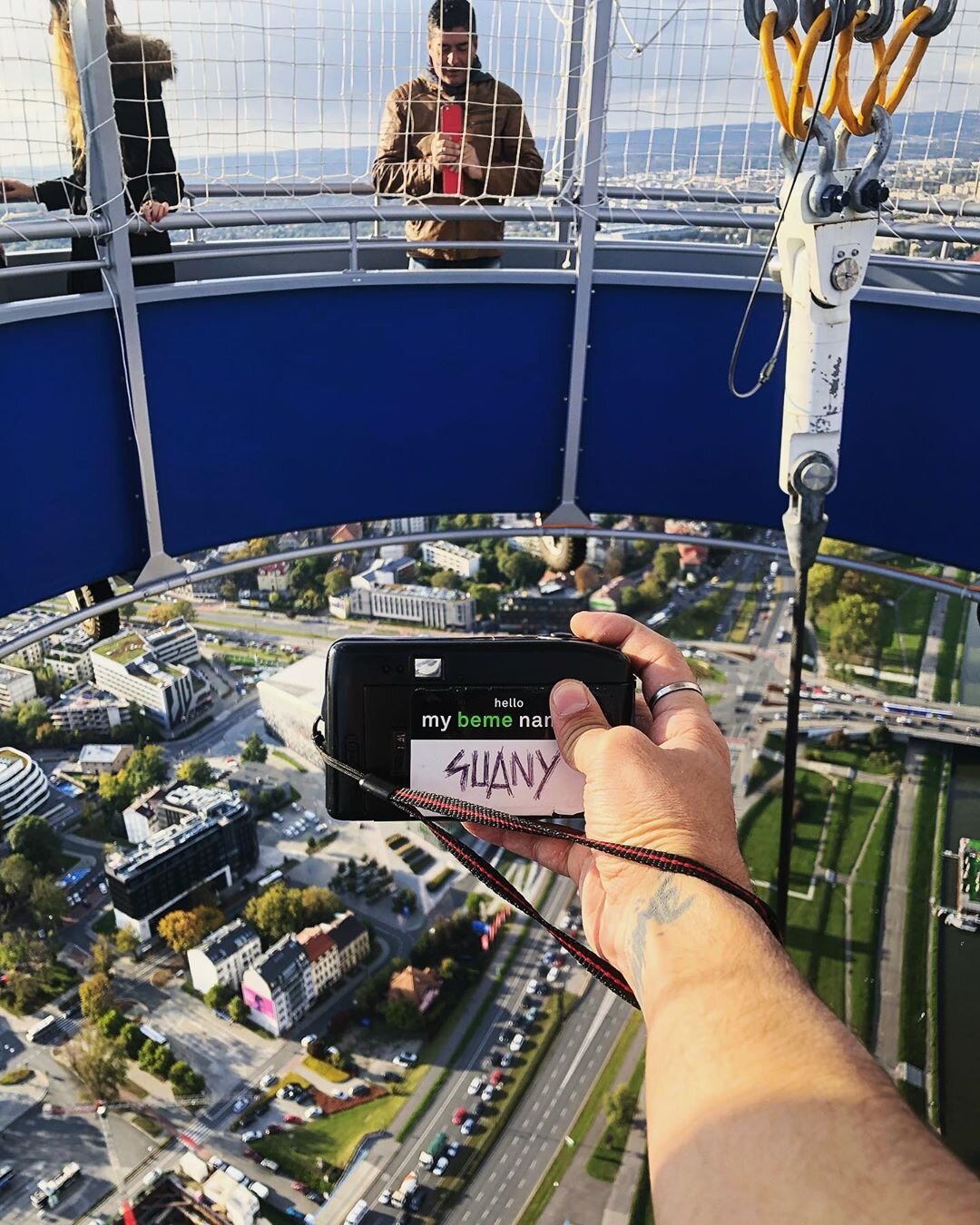 Person holding a camera with a note that says 'SUNNY' on a high observation platform above a cityscape with tall buildings, roads, and greenery.