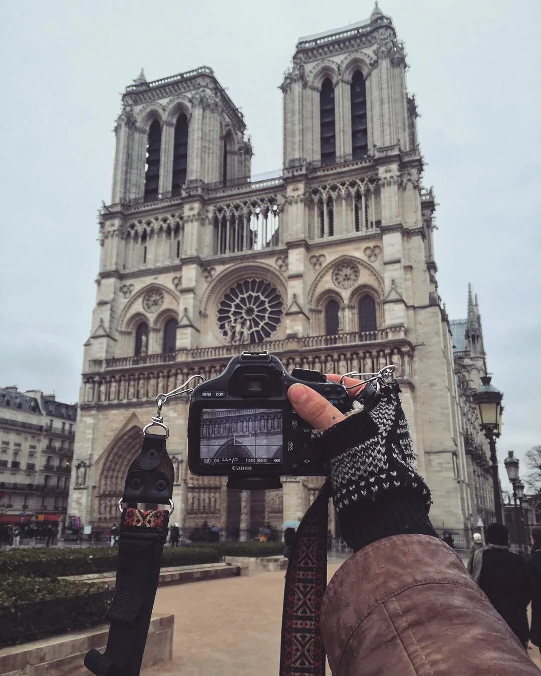 A person taking a photograph with a Canon camera of the Notre Dame Cathedral in Paris on a cloudy day. The person is wearing a brown coat and black-and-white patterned gloves. The cathedral's facade with twin towers and large rose window is visible i