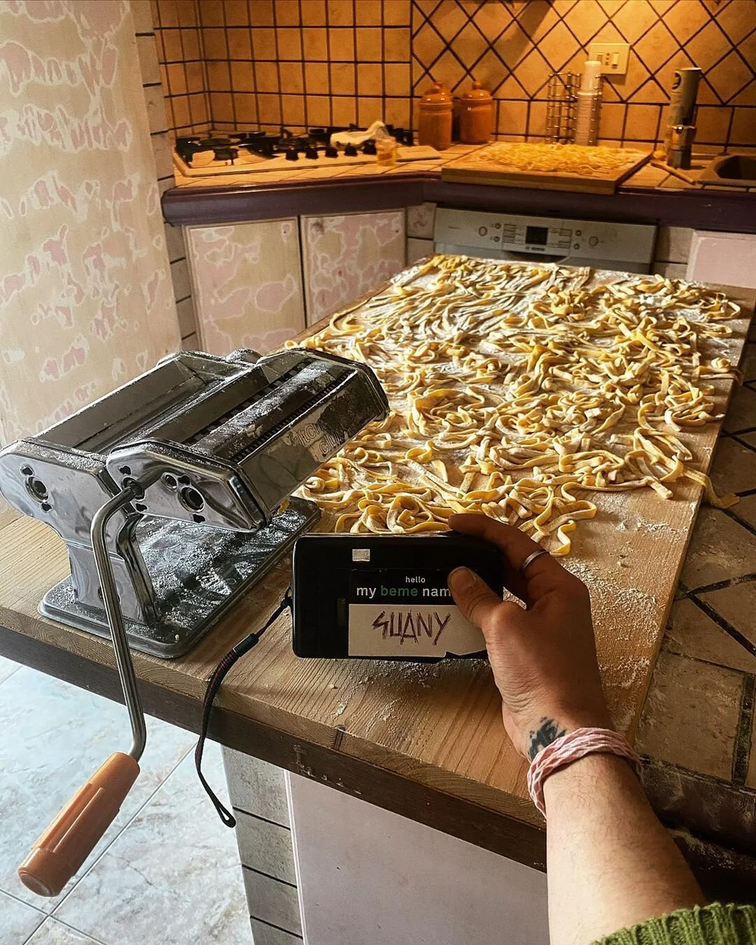 Unfinished homemade pasta spread on a wooden kitchen countertop with a pasta machine and a person holding a phone with a label reading 'SLADY' in the foreground.